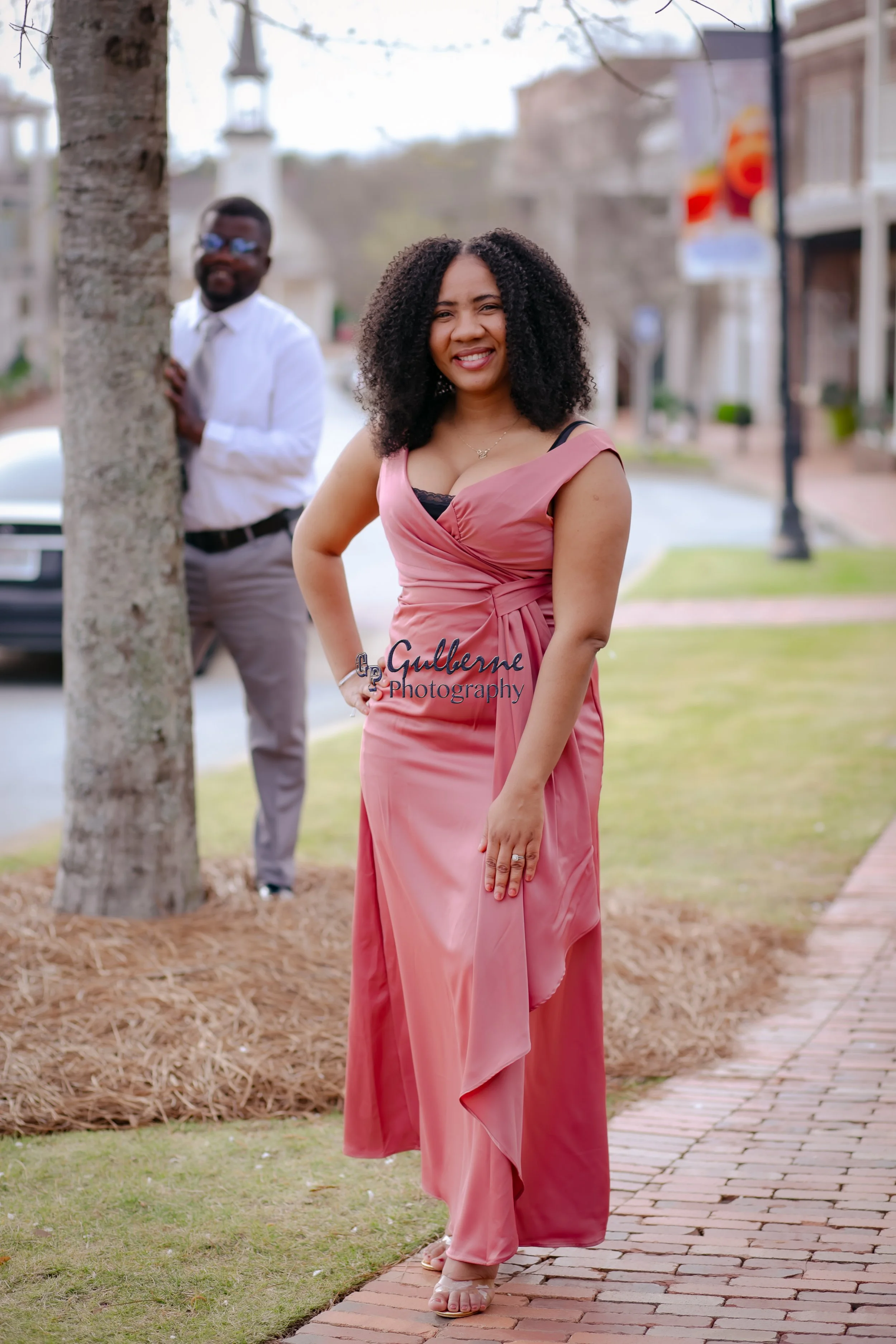 A woman in a pink dress standing outdoors on a brick pathway with a man in the background, partially hidden by a tree, wearing a white shirt and gray pants.