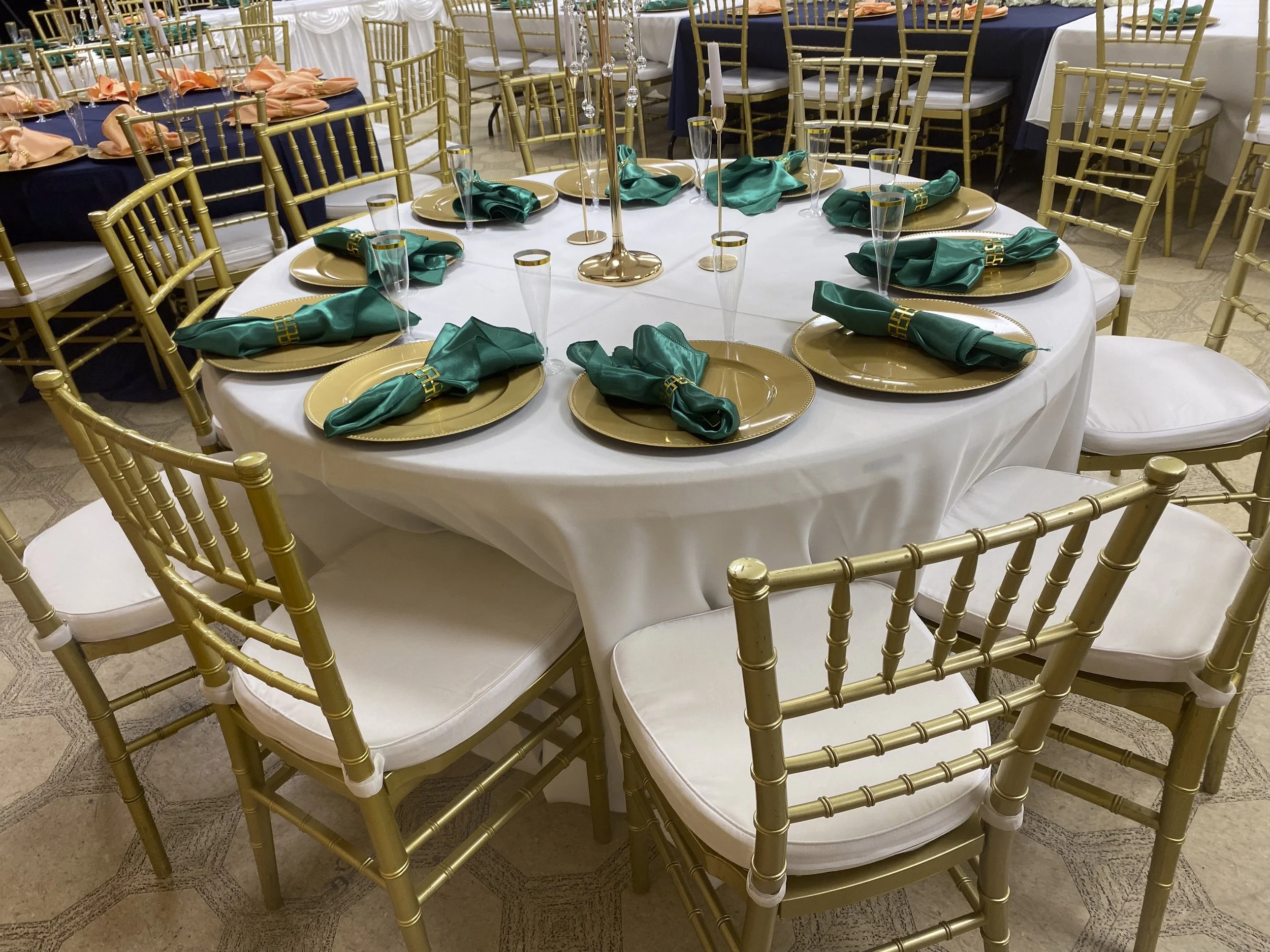A round banquet table covered with a white tablecloth, set with gold charger plates, green napkins, and clear champagne glasses, surrounded by gold Chiavari chairs, with candles and table decorations in the background.