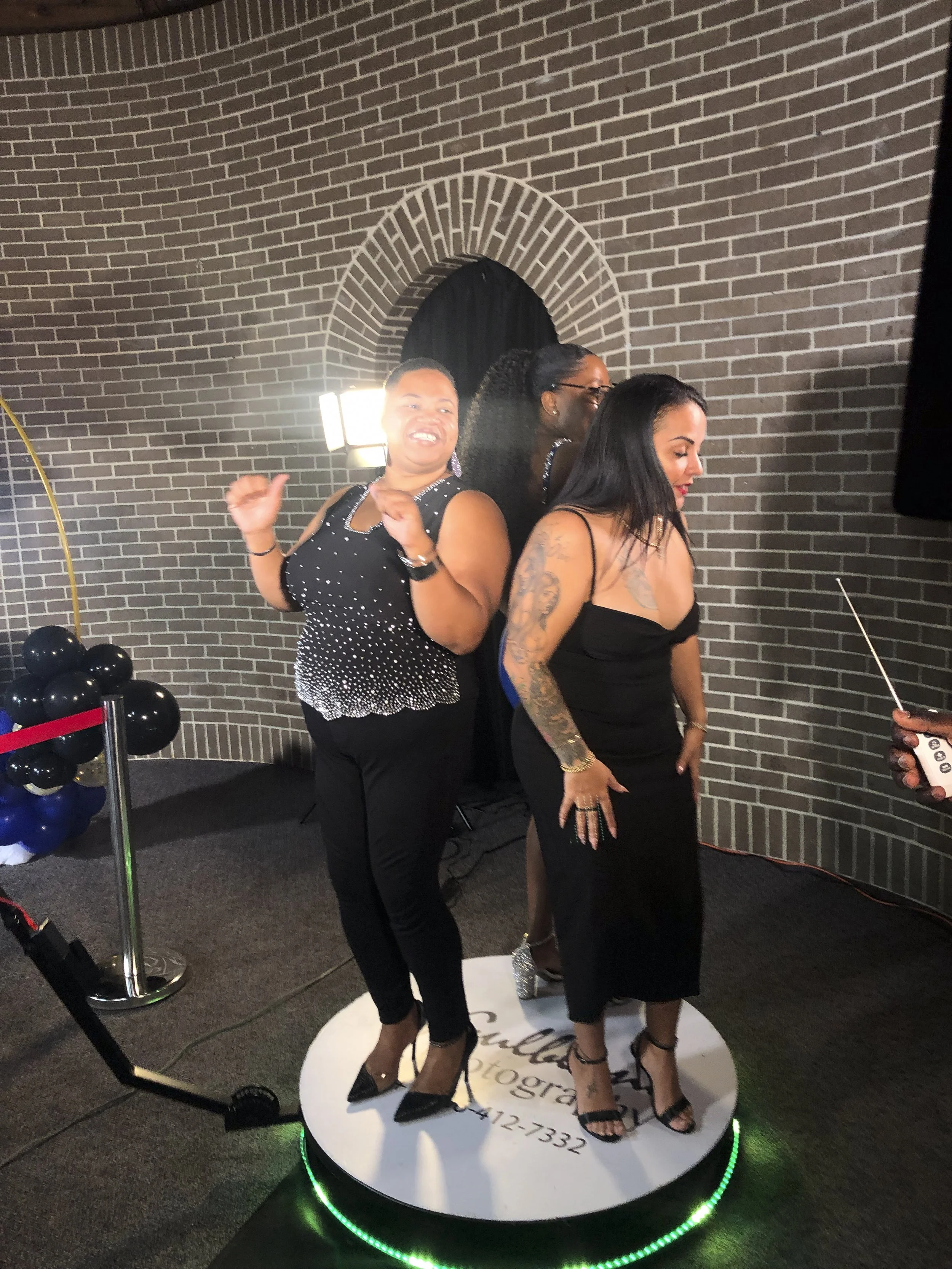 Three women are standing on a round platform with a sign for a photography studio. The platform has a green-lit edge. Two women are smiling and looking at the camera, while the third woman is looking down. There are black and blue balloons and a yell