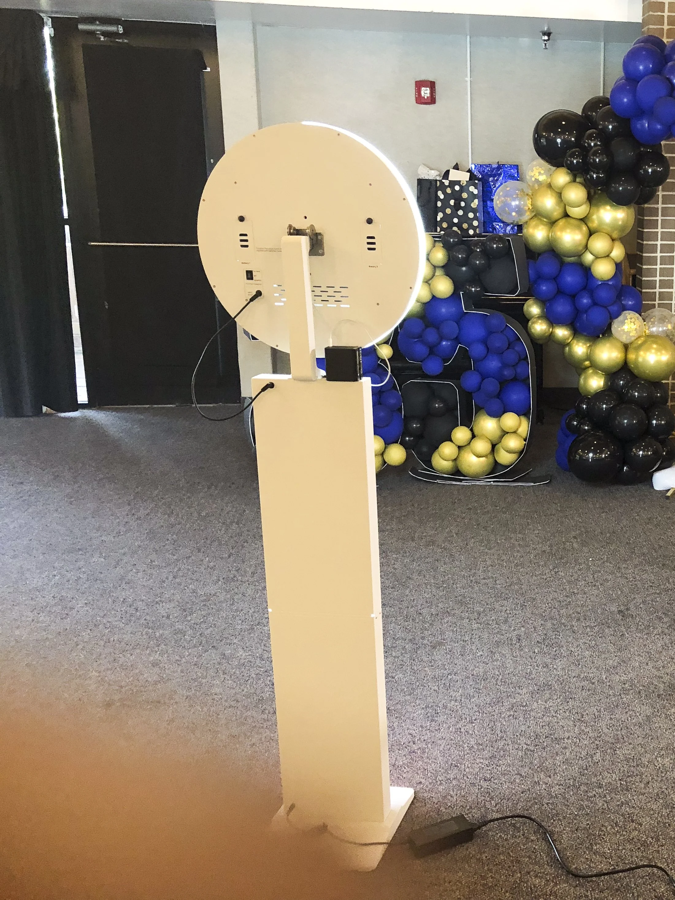 Photo of a photo booth prop in a corner, with a large circular photo prop on a stand, decorated with black, gold, and blue balloons arranged around it, and gift bags in the background.