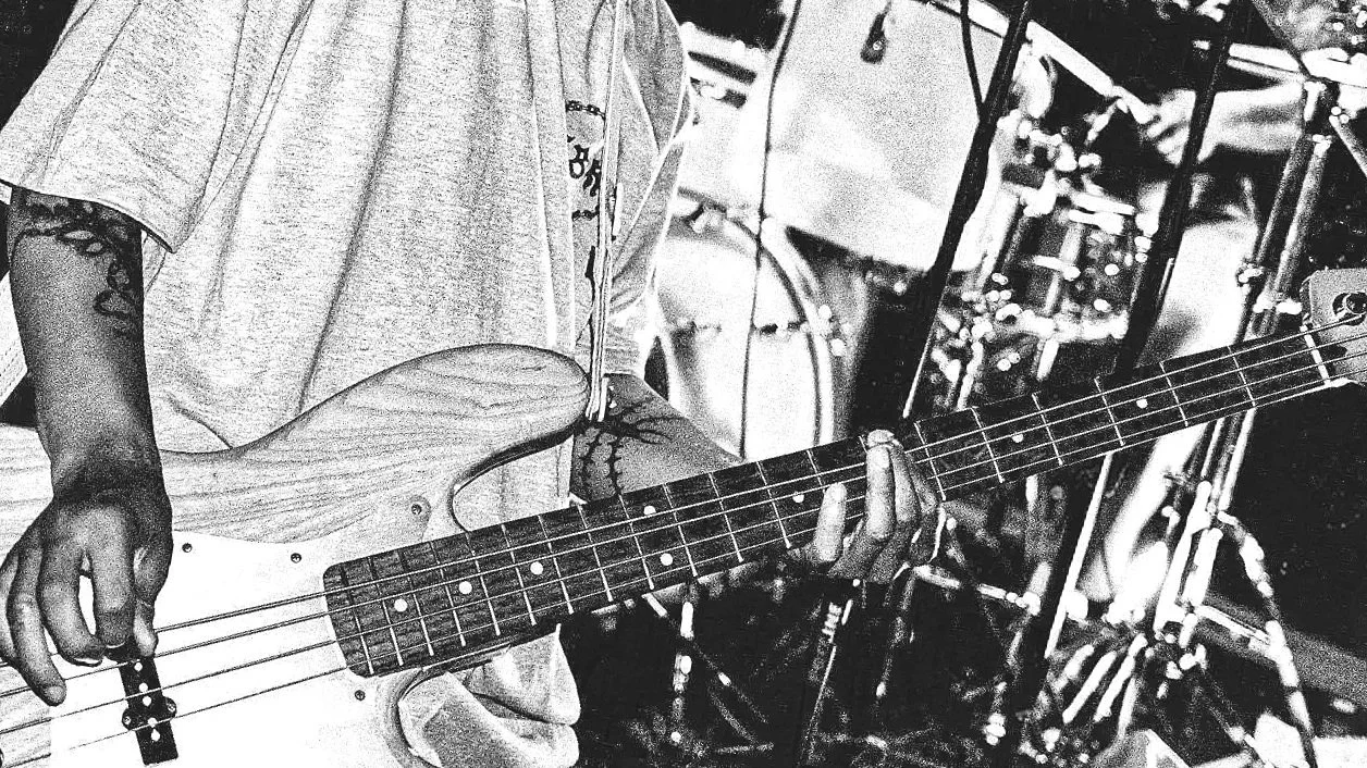 Close-up of a person playing an electric bass guitar in a music studio, with drums and other instruments in the background.