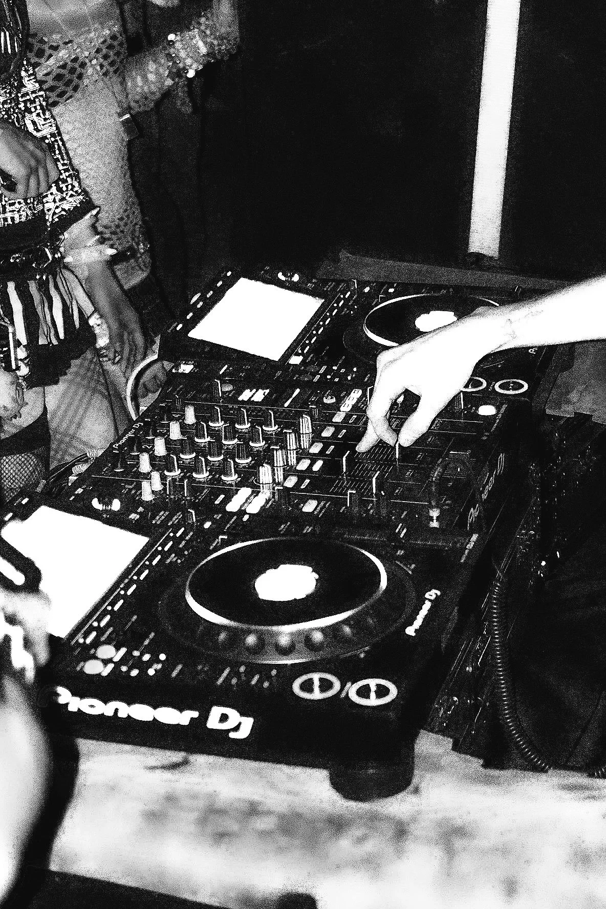 A DJ's setup with Pioneer DJ equipment, including a mixer and turntables, with a hand adjusting controls. Part of a person's arm is visible, and there are various hanging accessories in the background. The photo is in black and white.