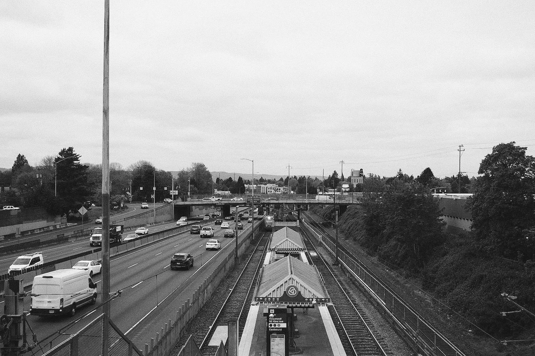 Black and white photo of a busy urban highway with multiple cars, a railway track with a train, and buildings in the background under a cloudy sky.
