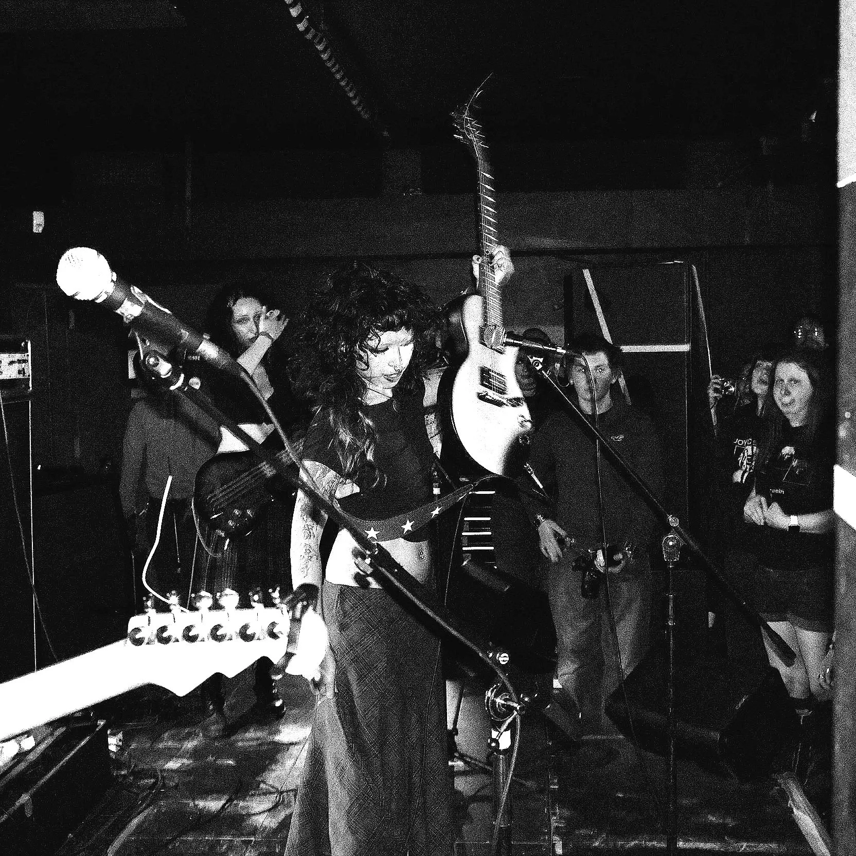 Black and white photo of a woman with curly hair singing into a microphone, holding a guitar, on a stage with other musicians and instruments around her.