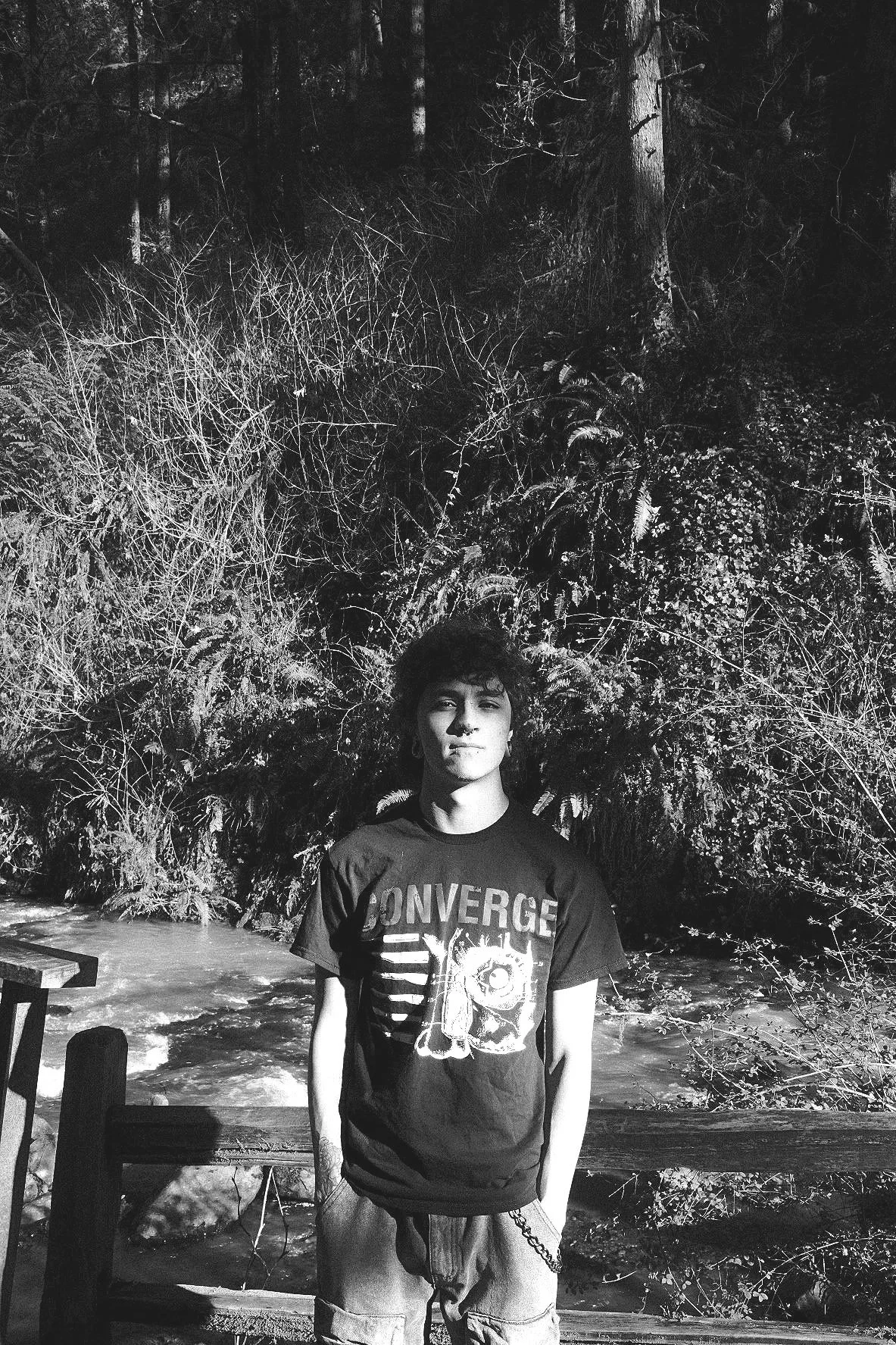 A young man with dark, curly hair stands in front of a wooded area with trees and foliage, beside a river or stream, wearing a T-shirt with the text 'Converge' and a graphic, and light-colored shorts.