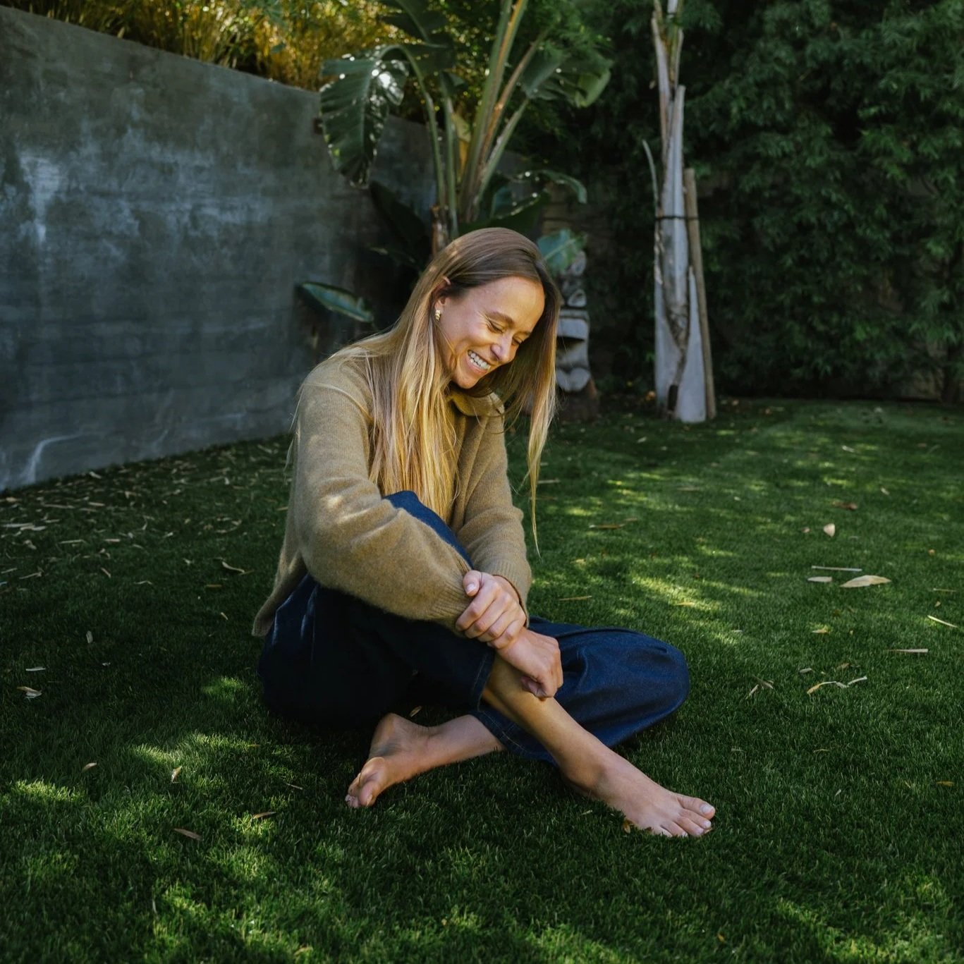 A woman with long blonde hair sat on a green lawn, smiling and stretching her legs. She is dressed in a tan sweater and dark pants, with a serene outdoor background including trees and a concrete wall.