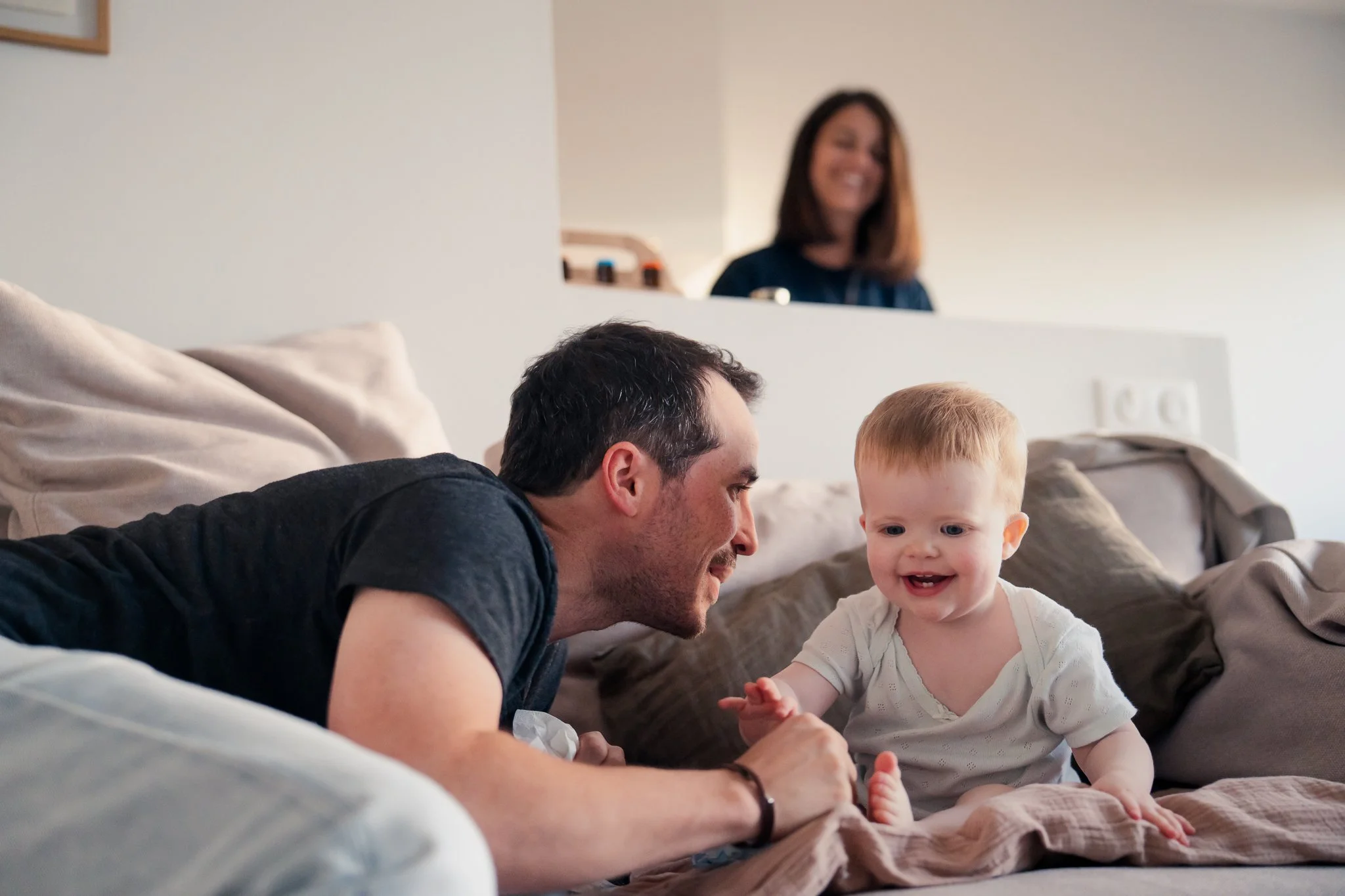 A man and a woman play with a young girl on a bed in a cozy bedroom. The man appears to be lying on his side, smiling at the girl, who has blonde hair and is wearing a white top. The woman is in the background, smiling and looking toward the scene.
