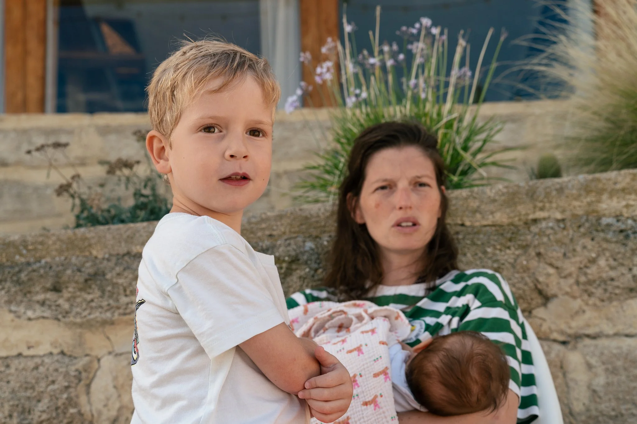 A young boy with blonde hair and a white t-shirt has his arm around a baby wrapped in a blanket, with a woman sitting behind them. They are outdoors near a stone wall with plants in the background.