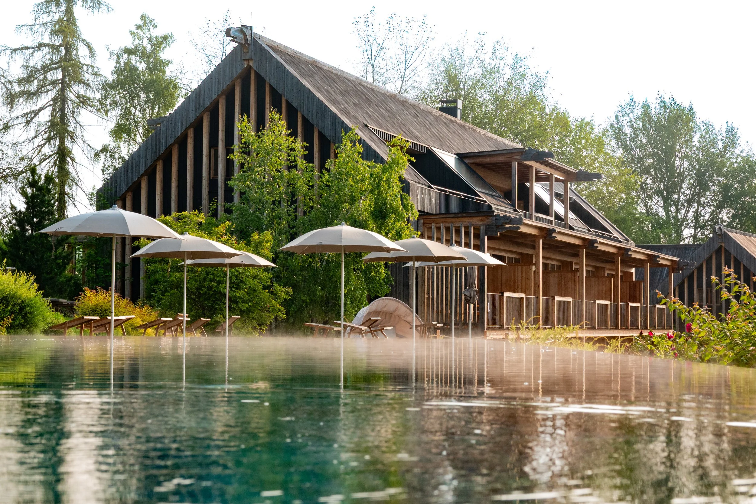 A rustic house with a steep sloped roof surrounded by trees, with outdoor lounge chairs and umbrellas near a steaming hot spring or pool.