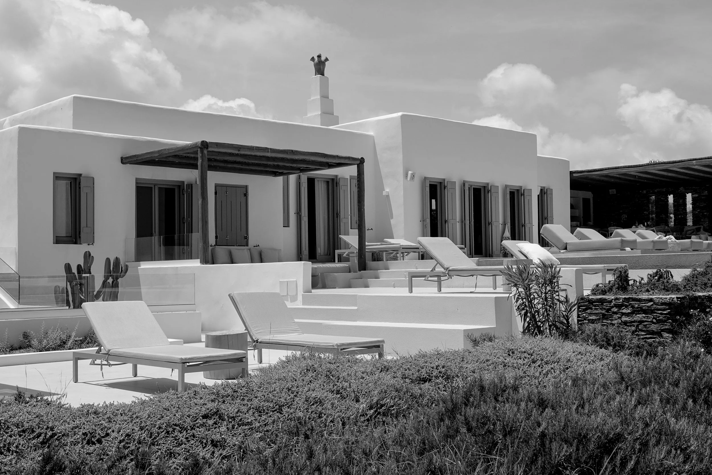 A black-and-white photo of a modern villa with multiple flat roofs, window shutters, and outdoor lounging areas, with sun loungers and a garden with various plants in the foreground.
