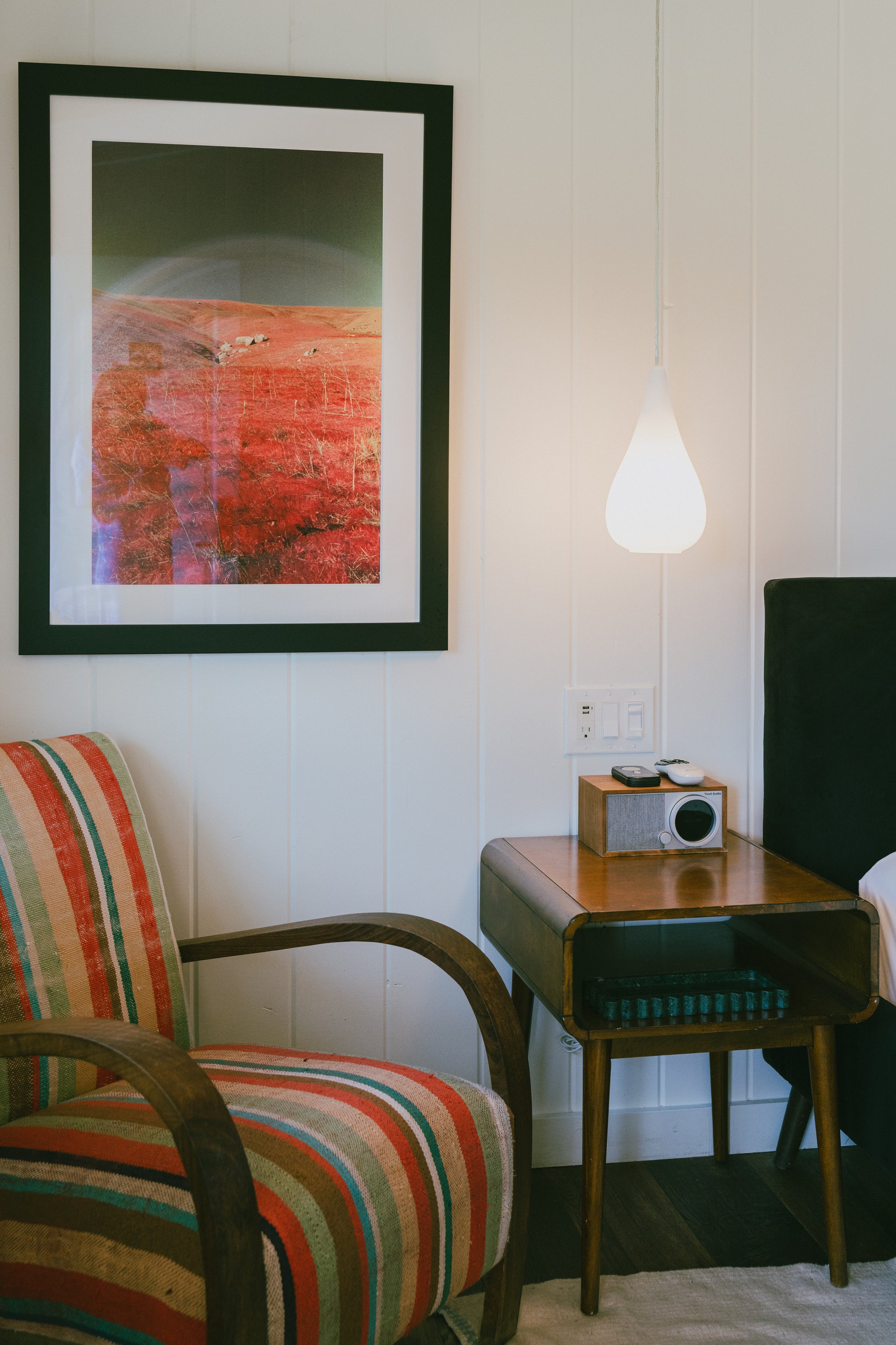 Living room with a framed landscape painting of a reddish-orange desert on the wall, a modern white teardrop-shaped pendant light, a wooden side table with a radio and remote, an armchair with multicolored striped fabric, and a black upholstered chai