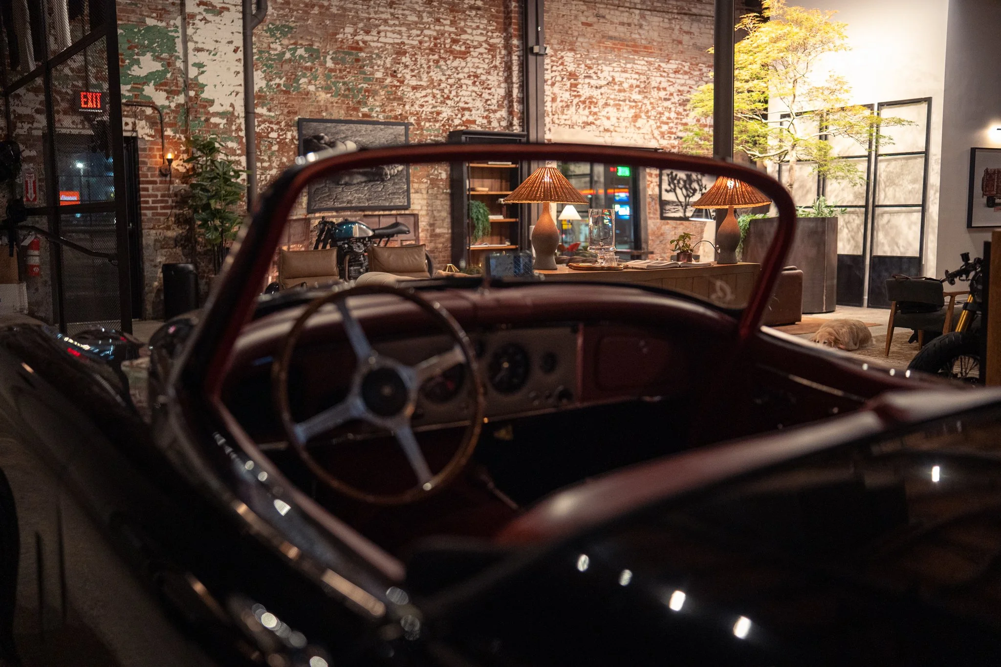 Interior of a cozy, industrial-style living space with exposed brick walls, large windows, and warm lighting. There is a vintage car dashboard in the foreground, and furniture including a brown sofa, lamps, and framed artwork. A cat is sleeping on the floor near the furniture.