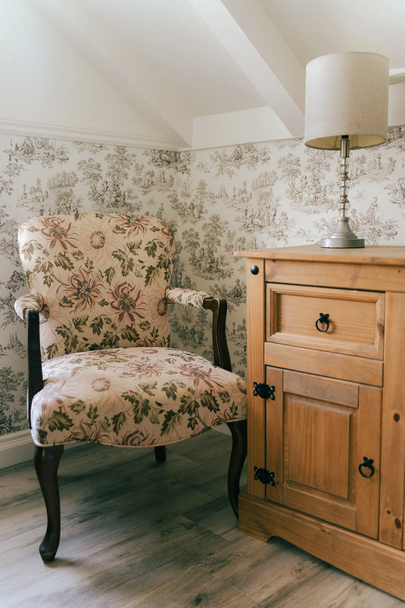 A vintage upholstered armchair with floral pattern next to a wooden cabinet with a lampshade on top, set against wallpaper with toile de Jouy pattern.