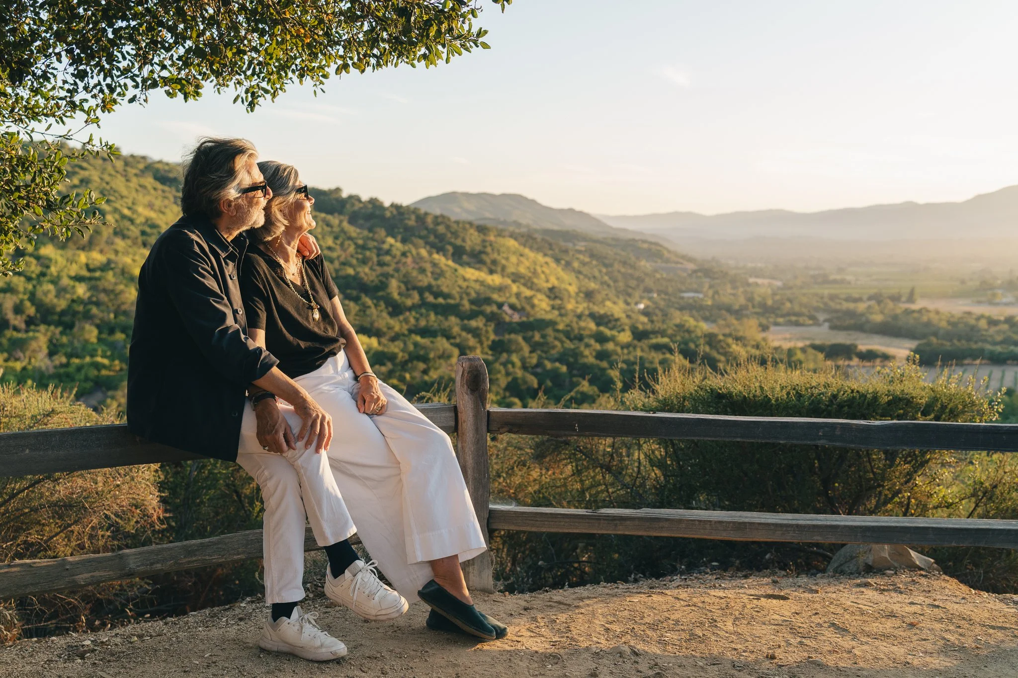 An elderly man and woman sitting together on a wooden fence overlooking a landscape of rolling hills, trees, and a bright sky, enjoying a peaceful moment outdoors.