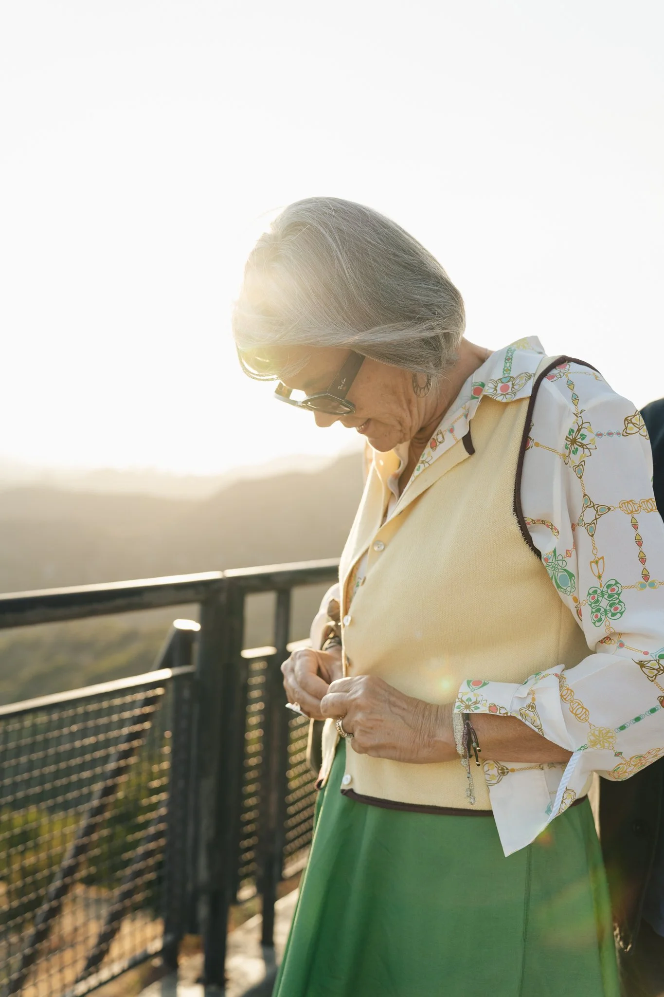 Older woman with gray hair wearing glasses, a yellow vest, a floral shirt, and a green skirt standing outdoors by a black metal fence, with sunlight shining behind her.