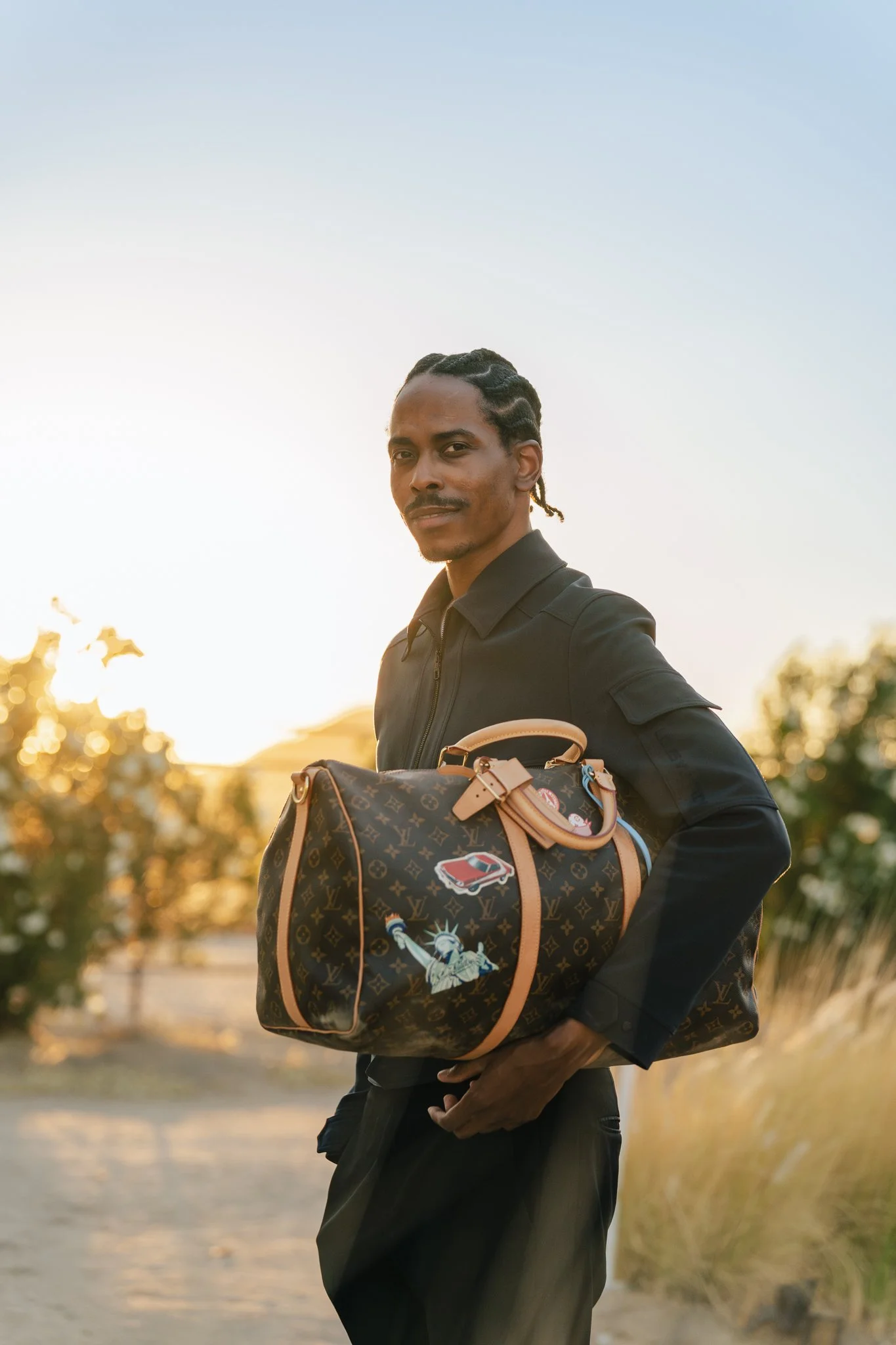 A man with braided hair and a mustache wearing a black jacket, holding a patterned Louis Vuitton bag with patches, standing outdoors at sunset.