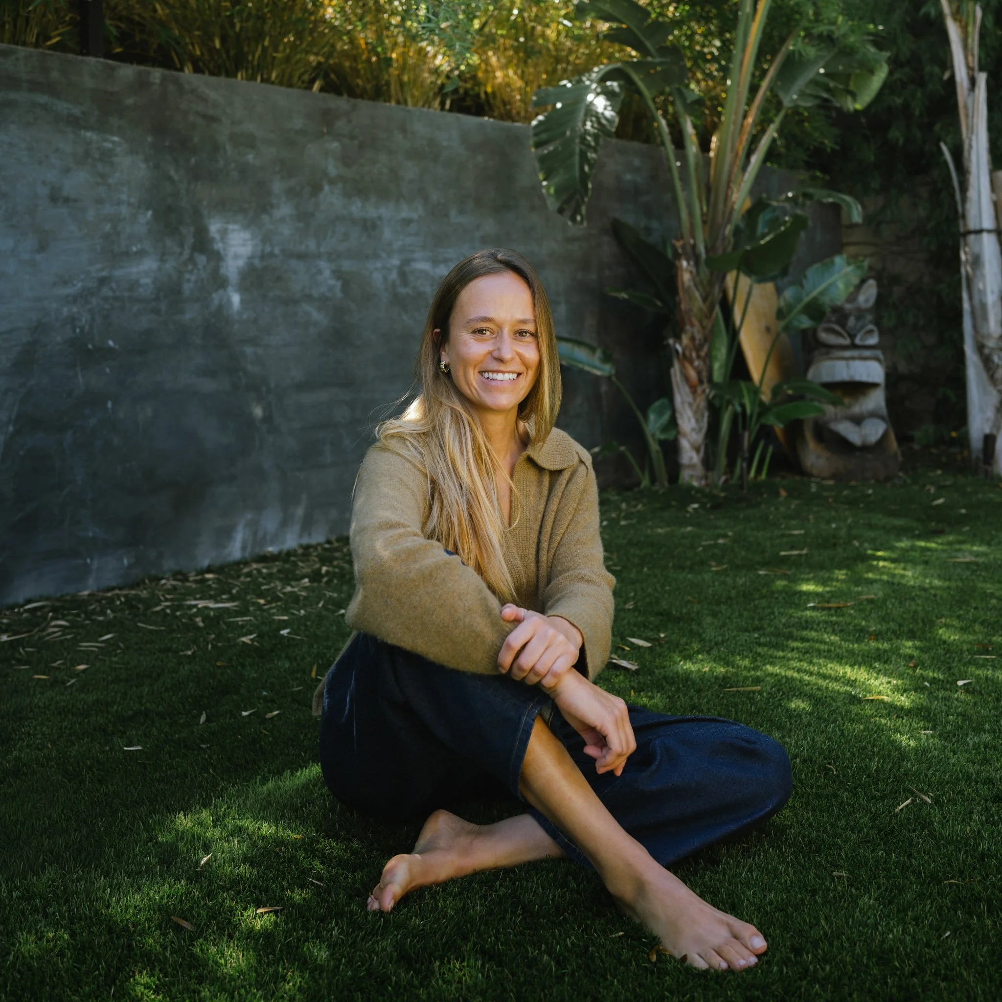 A woman with long blonde hair smiling outdoors, sitting on green grass, with plants and a concrete wall in the background.