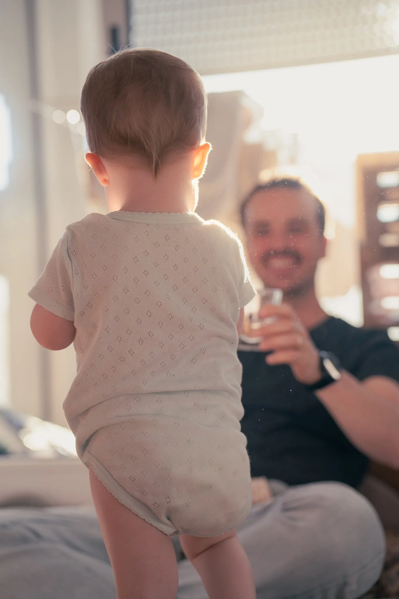 A toddler standing on a bed, facing away from the camera, with an adult woman smiling and holding a glass in the background. The sunlight is bright and streaming in from behind.