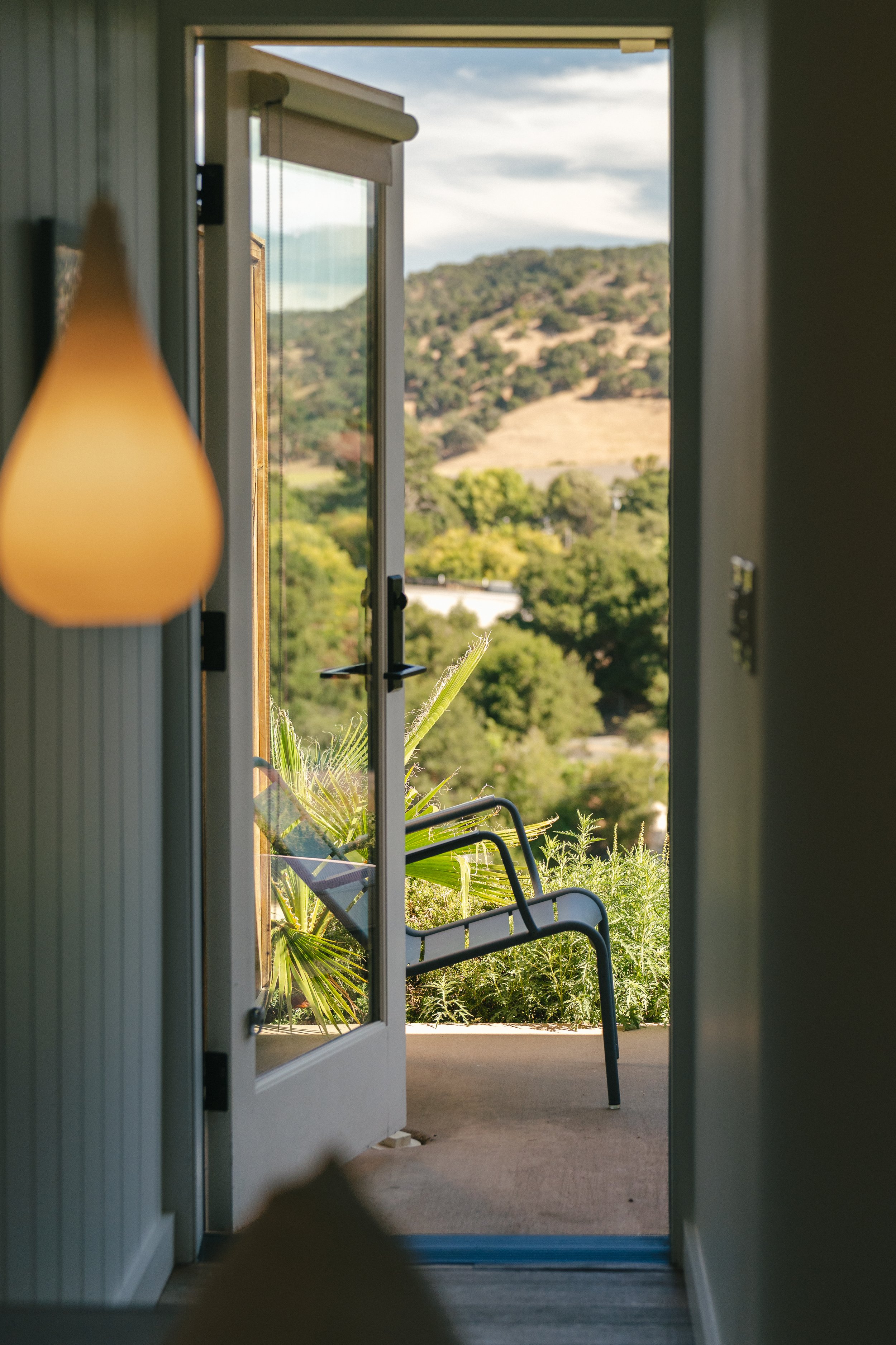 View through a partially open glass door showing outdoor chairs, plants, and a hillside with trees in the background.