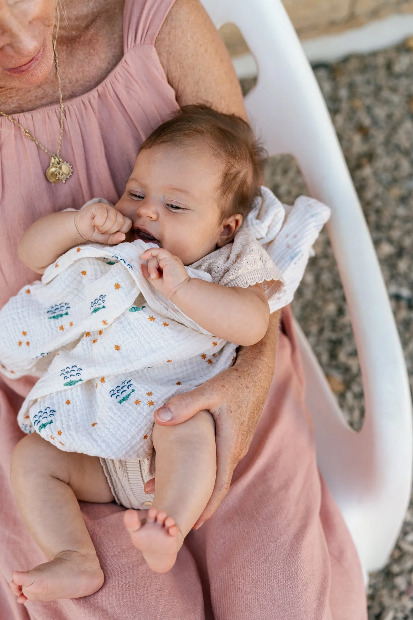 An elderly person holding a baby on their lap outdoors. The baby is wearing a patterned white cloth and is sucking on their fingers. The older person is wearing a pink dress with a gold necklace. The background shows a white chair and gravel ground.
