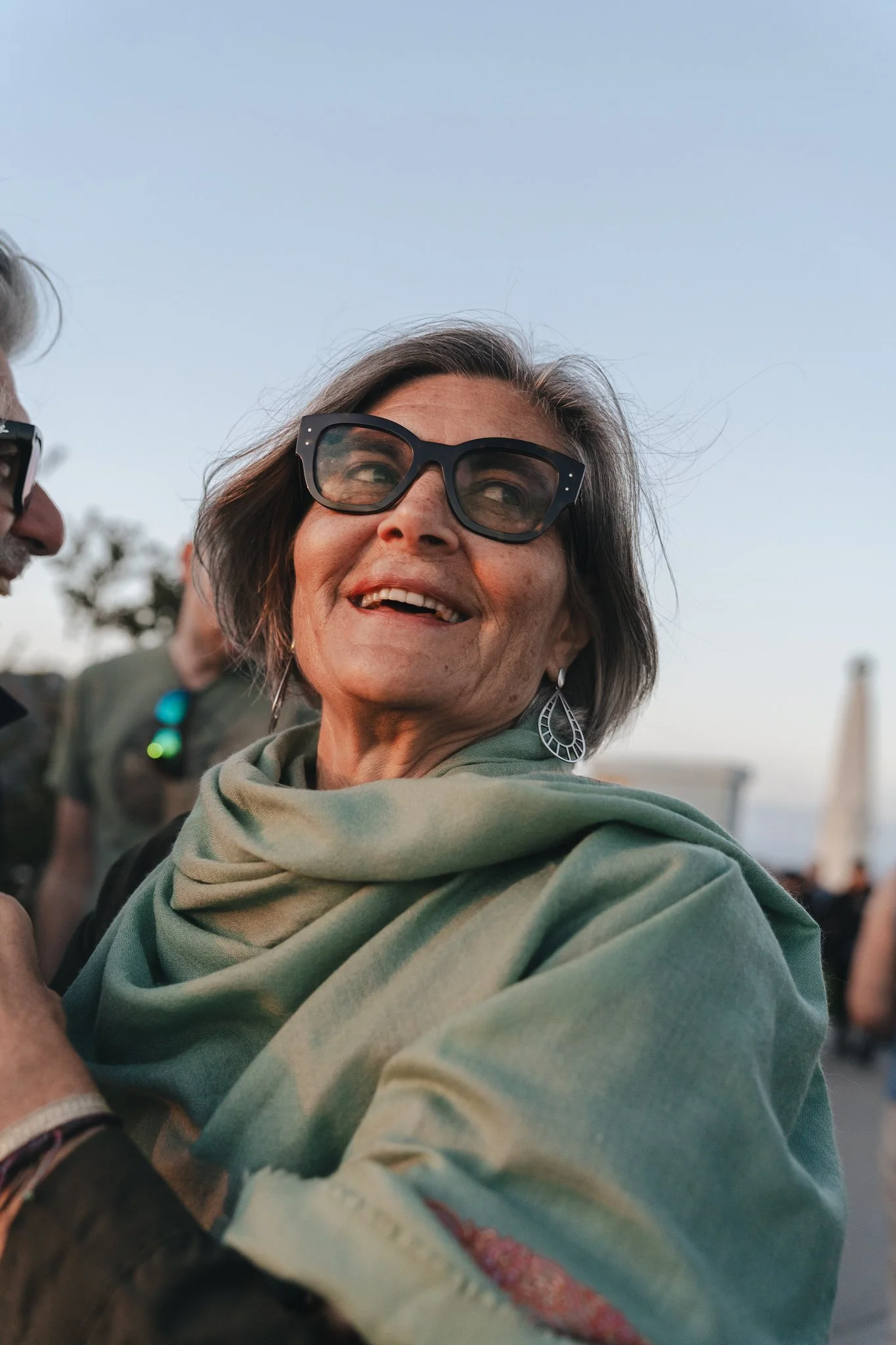 Close-up of an older woman smiling, wearing large black sunglasses and a green scarf, in an outdoor setting with a clear sky in the background.