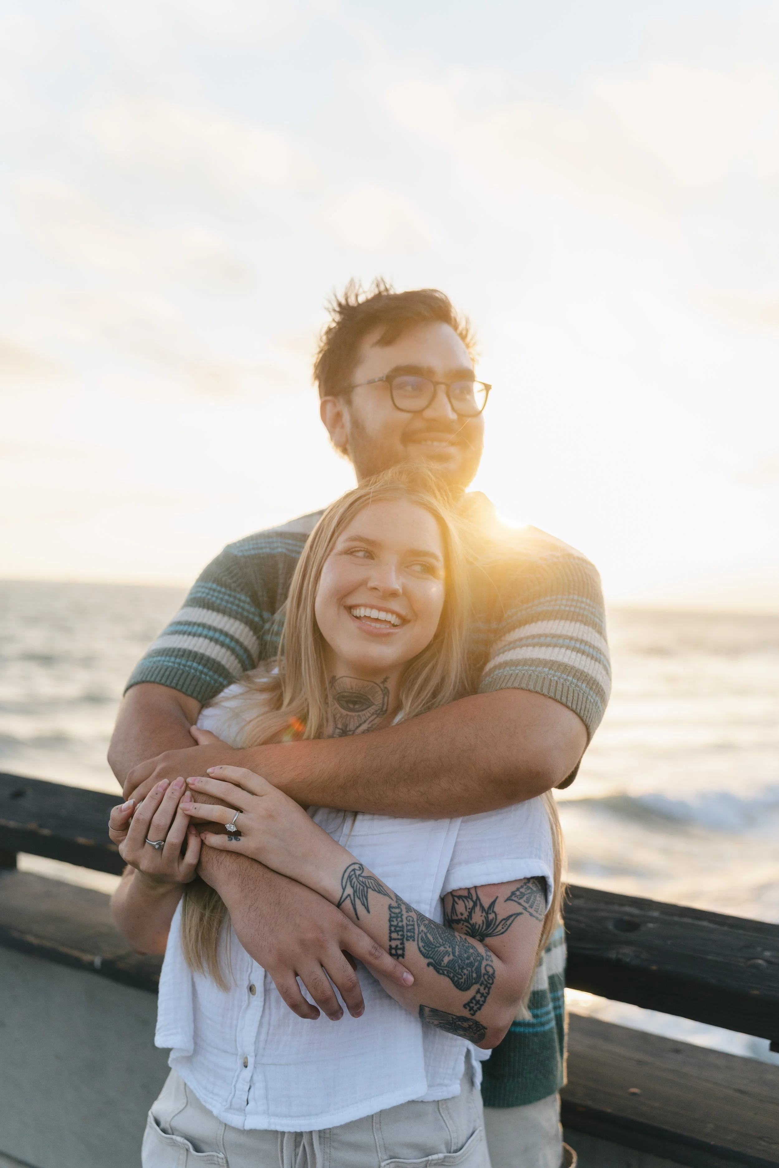 A couple standing on a wooden dock by the ocean during sunset, embracing and smiling, with tattoos on the woman's arms and chest, and the man wearing glasses.
