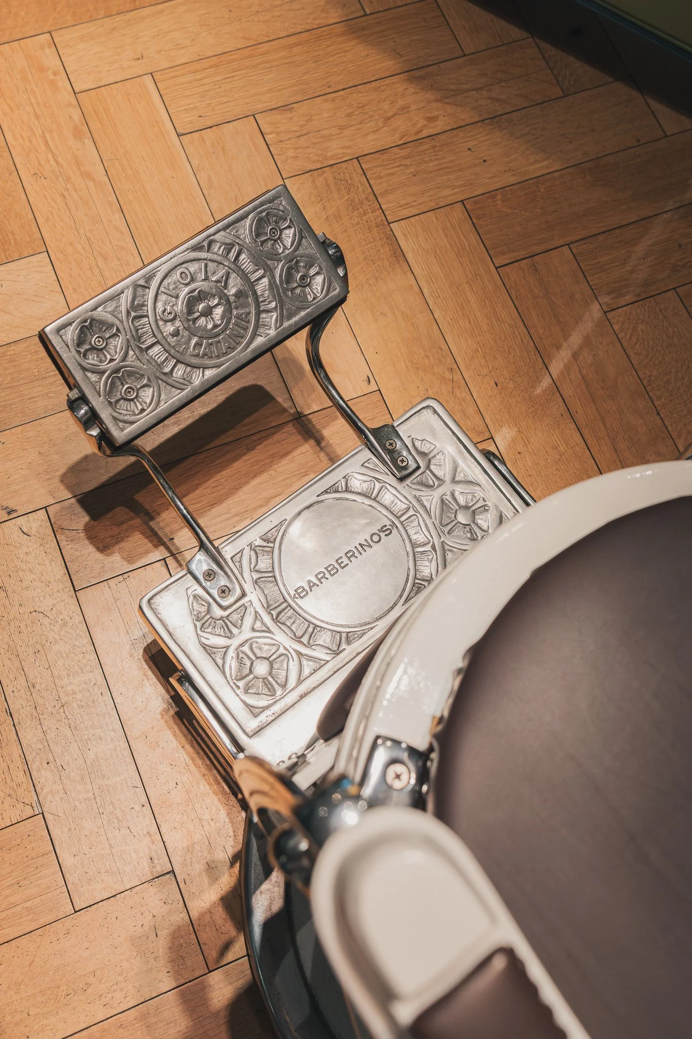 Close-up of a metallic footrest with decorative floral patterns and the word "ARBERINOS" inscribed, situated on a wooden floor, with part of a white toilet visible in the bottom right corner.
