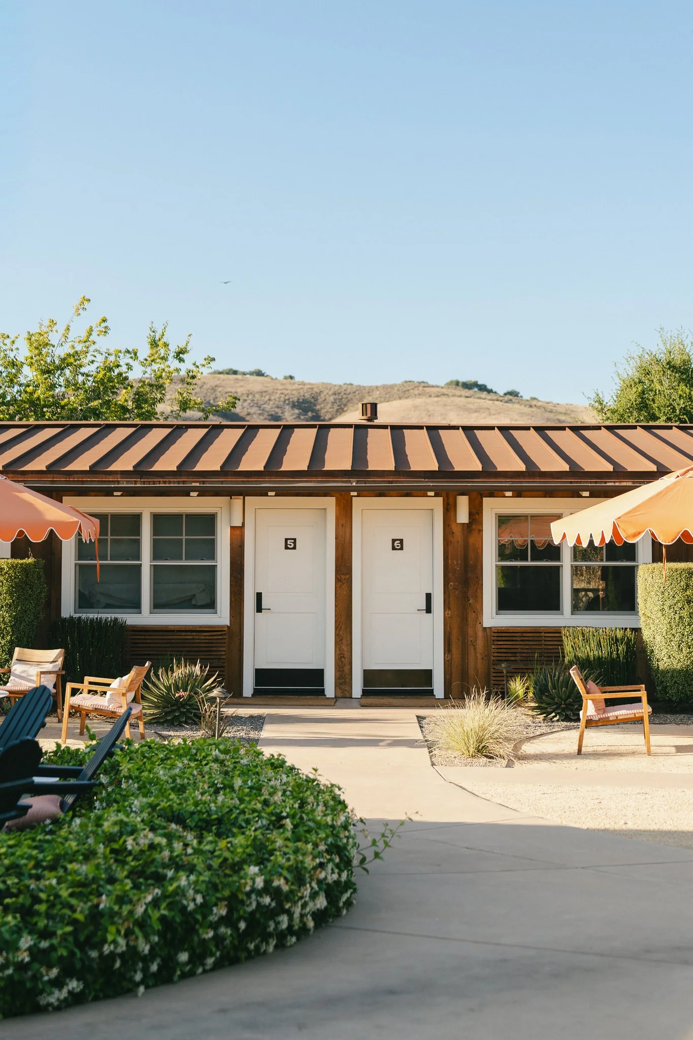 A building with two white doors numbered 5 and 6, a metal roof, surrounded by outdoor furniture, green bushes, and desert landscape with hills in the background under a clear blue sky.