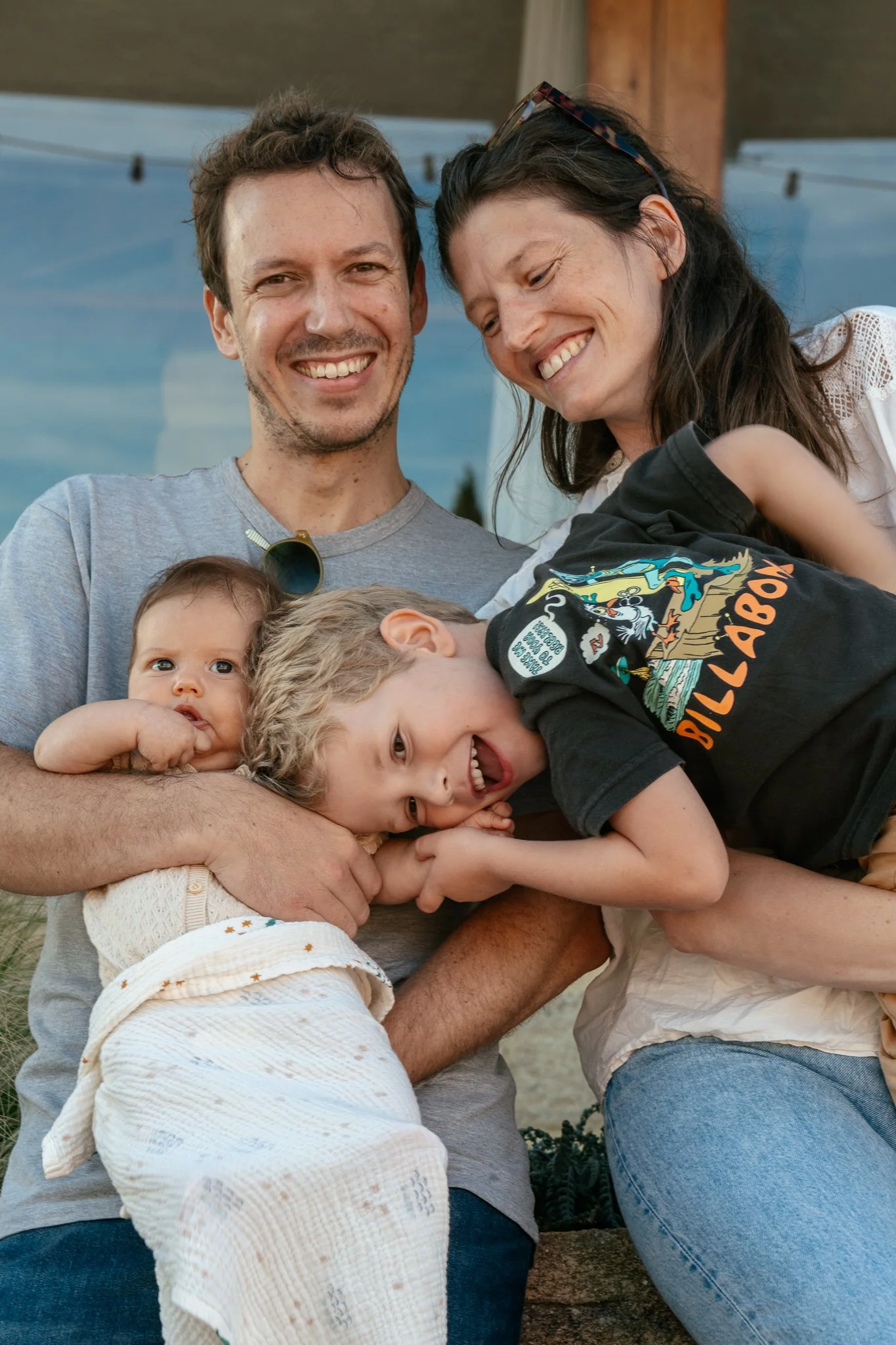 A smiling man holding two young children in an outdoor setting. The children are cuddled close, with one of them resting her head on the man's arm and the other smiling happily. The woman next to them is also smiling, wearing sunglasses on her head.