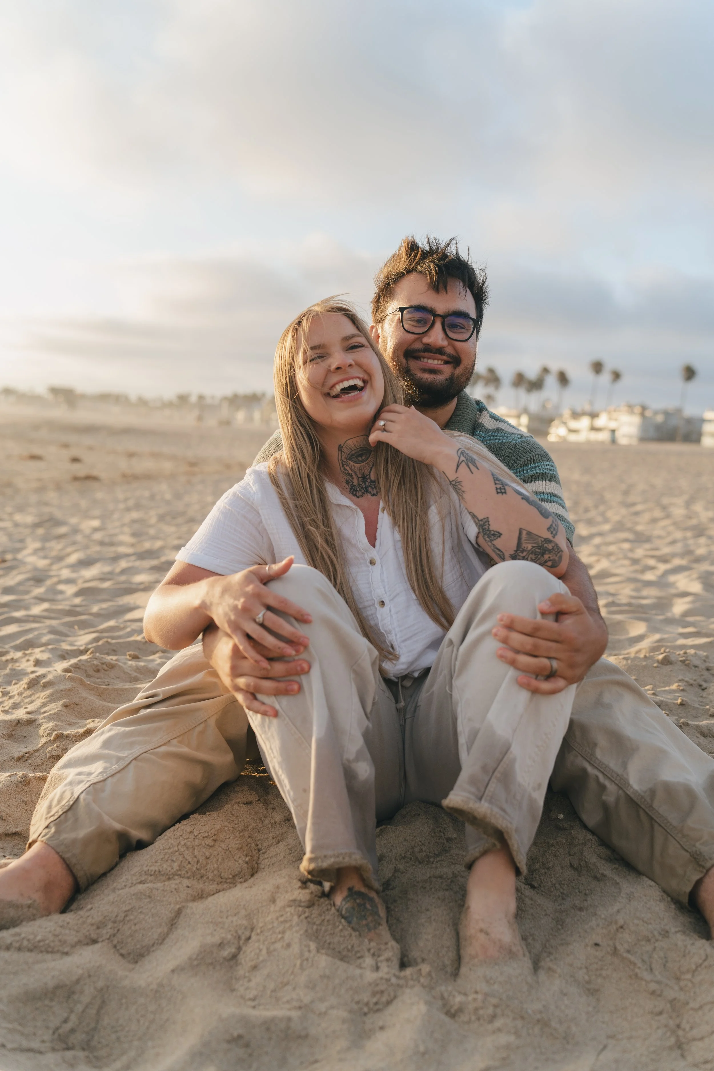 A smiling couple with tattoos sitting on the sand at the beach, with palm trees and buildings in the background under a cloudy sky.