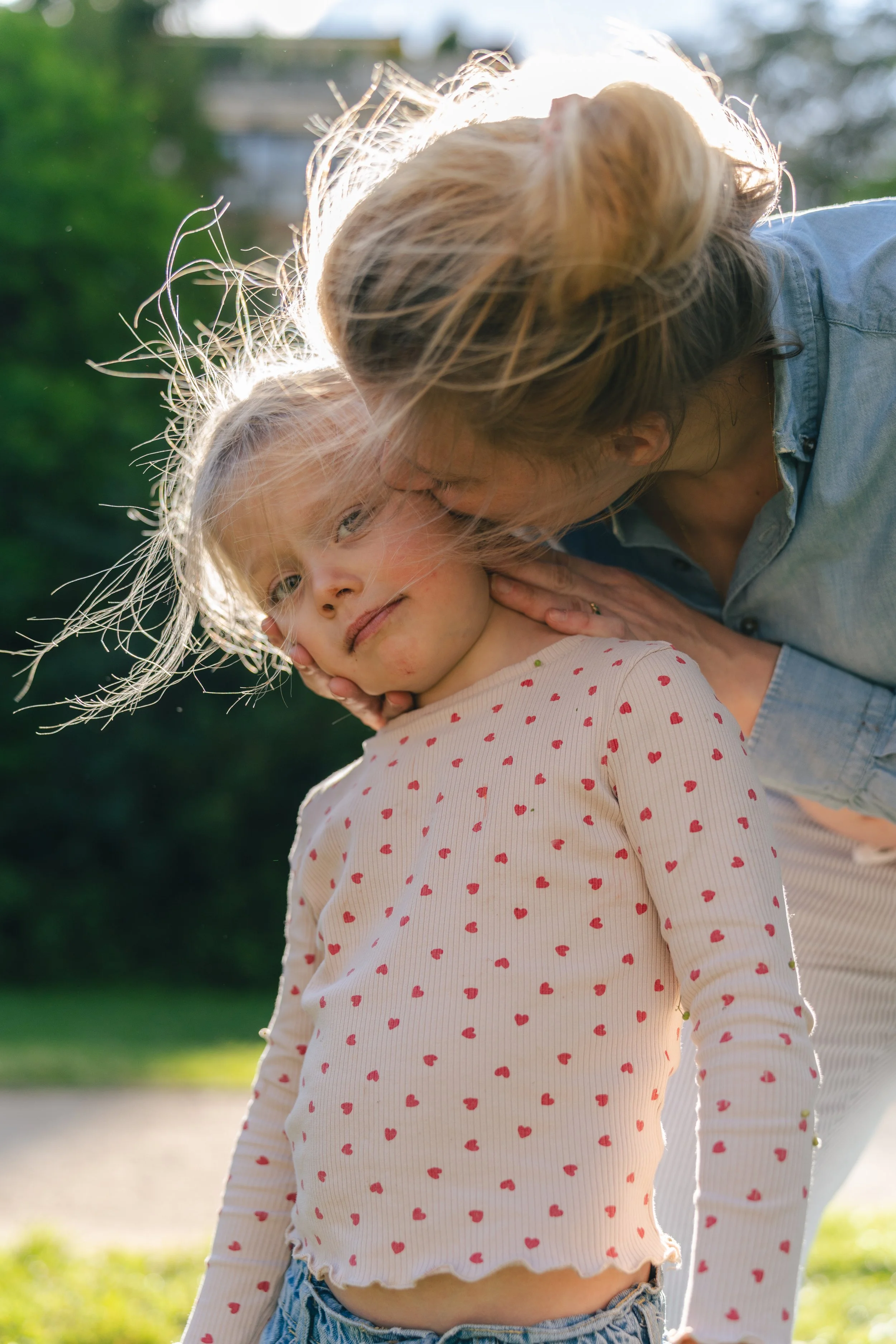 A woman gently kisses a young girl on the head outdoors during sunset. The girl wears a cream-colored shirt with red hearts and has blonde hair, while the woman has light brown hair and is dressed in a light blue shirt. The background displays blurre