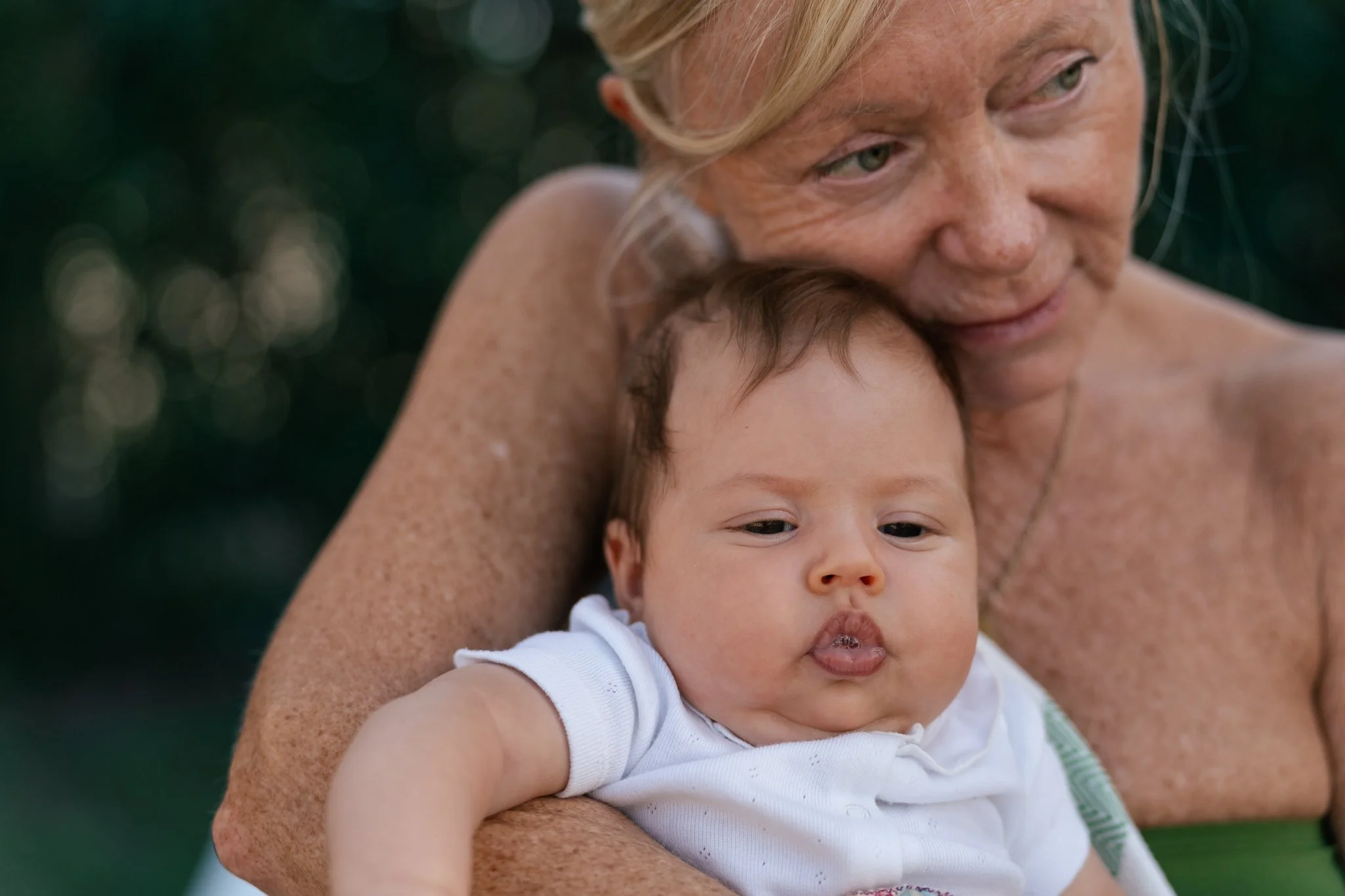 An older woman with blonde hair and freckles holding a young child with short brown hair, both outdoors. The woman is gently resting her face on the child's head.