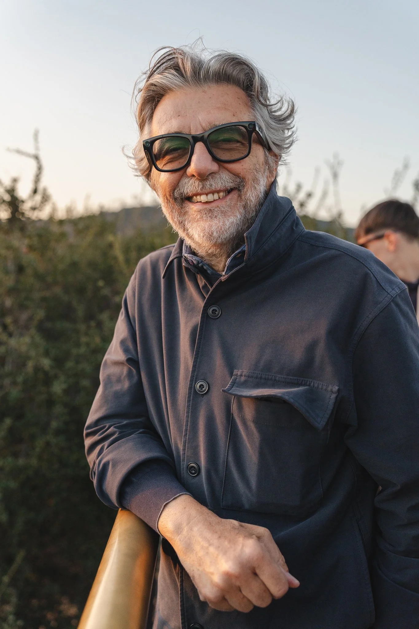 A smiling man with gray hair, beard, and large sunglasses, wearing a dark gray button-up shirt, outdoors with trees in the background.