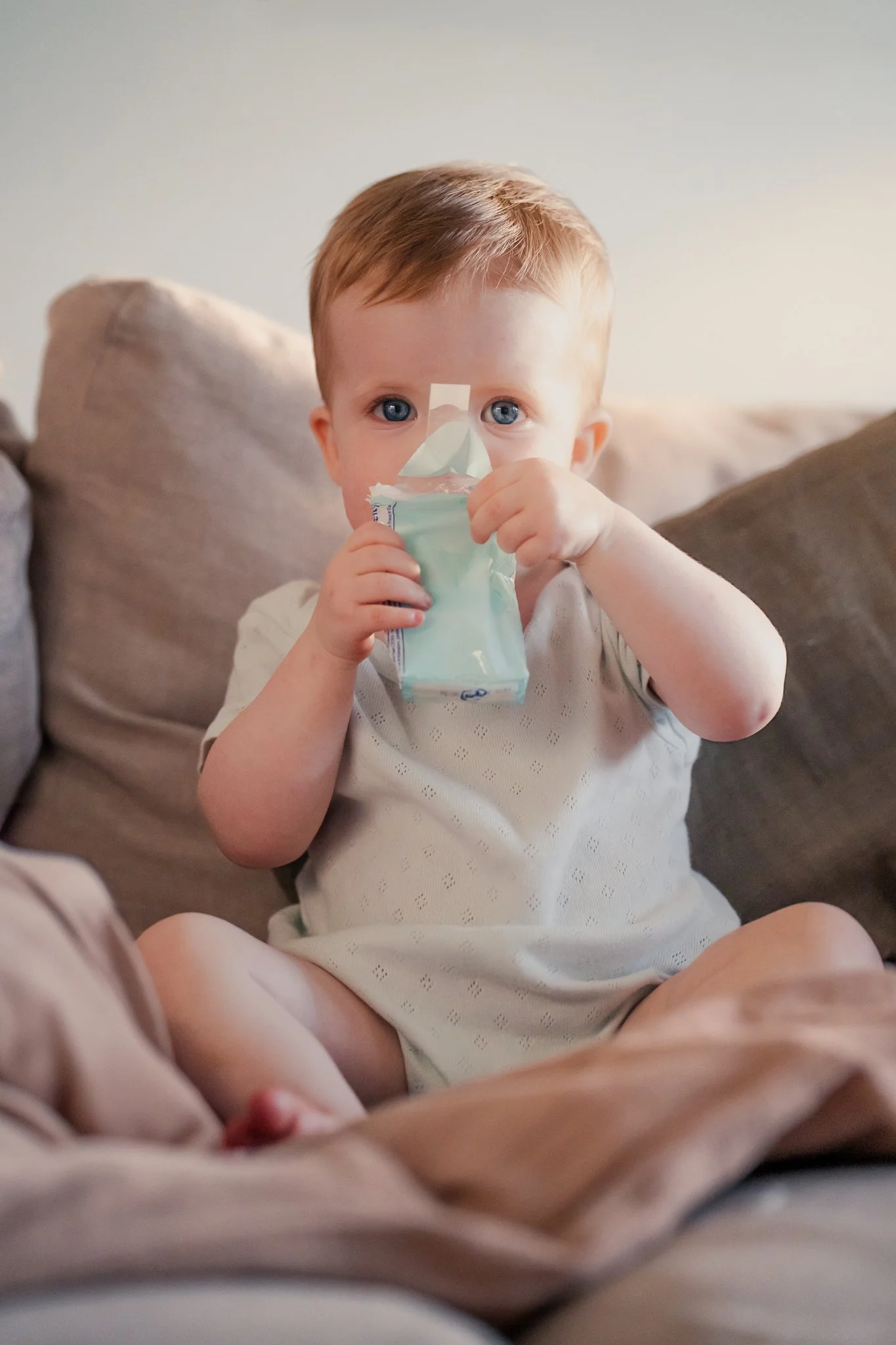 A young child with red hair and blue eyes sitting on a gray couch, holding and drinking from a juice pouch, with a plain light-colored wall in the background.