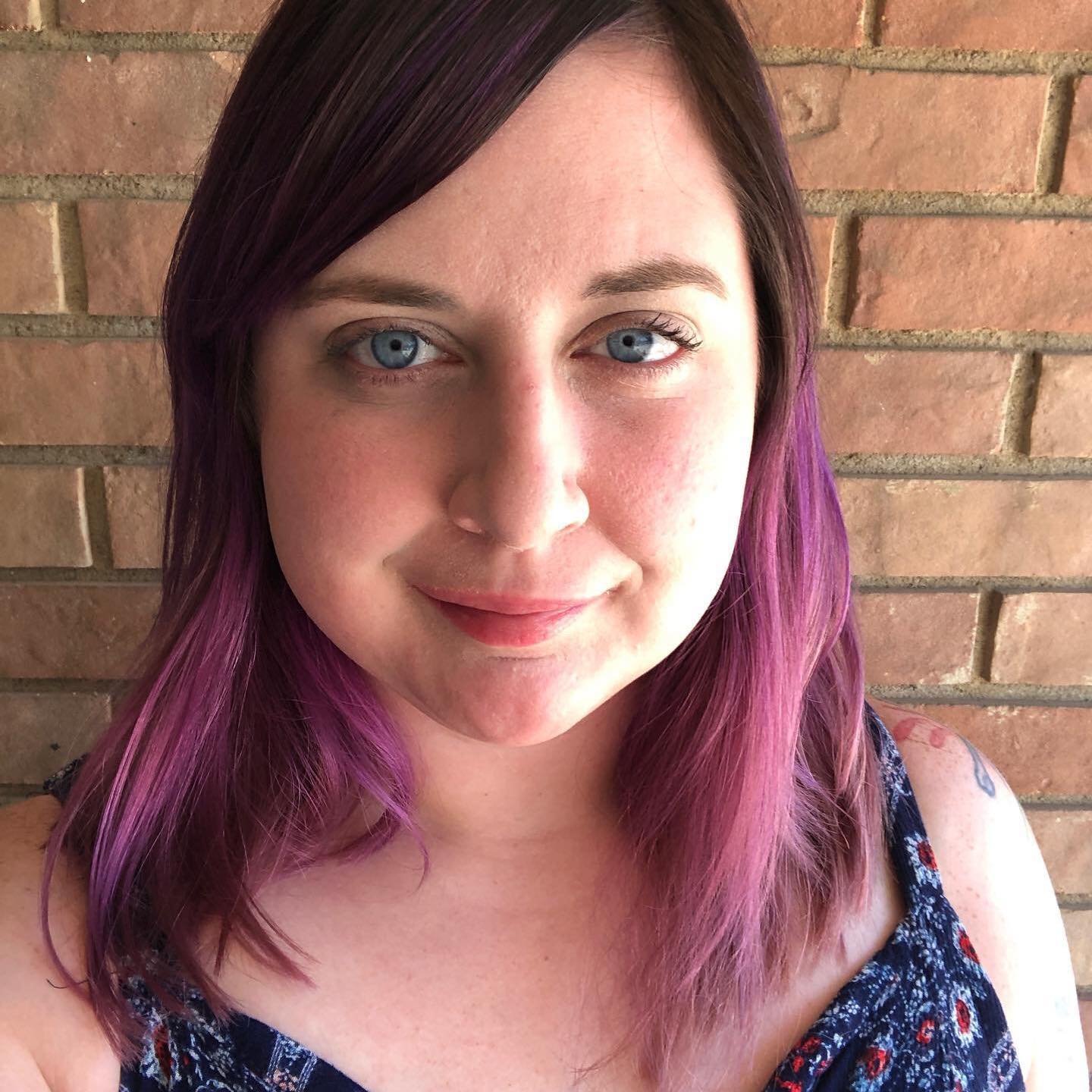 A woman with purple-tinted hair smiling in front of a brick wall