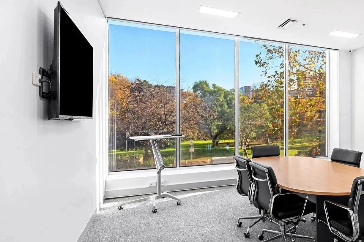 Flagstaff Mediation Centre - Flagstaff Mediation Room 1 with a large window overlooking Flagstaff Gardens with trees and autumn foliage. The room features a round wooden table surrounded by six black office chairs and a wall-mounted flat-screen TV.