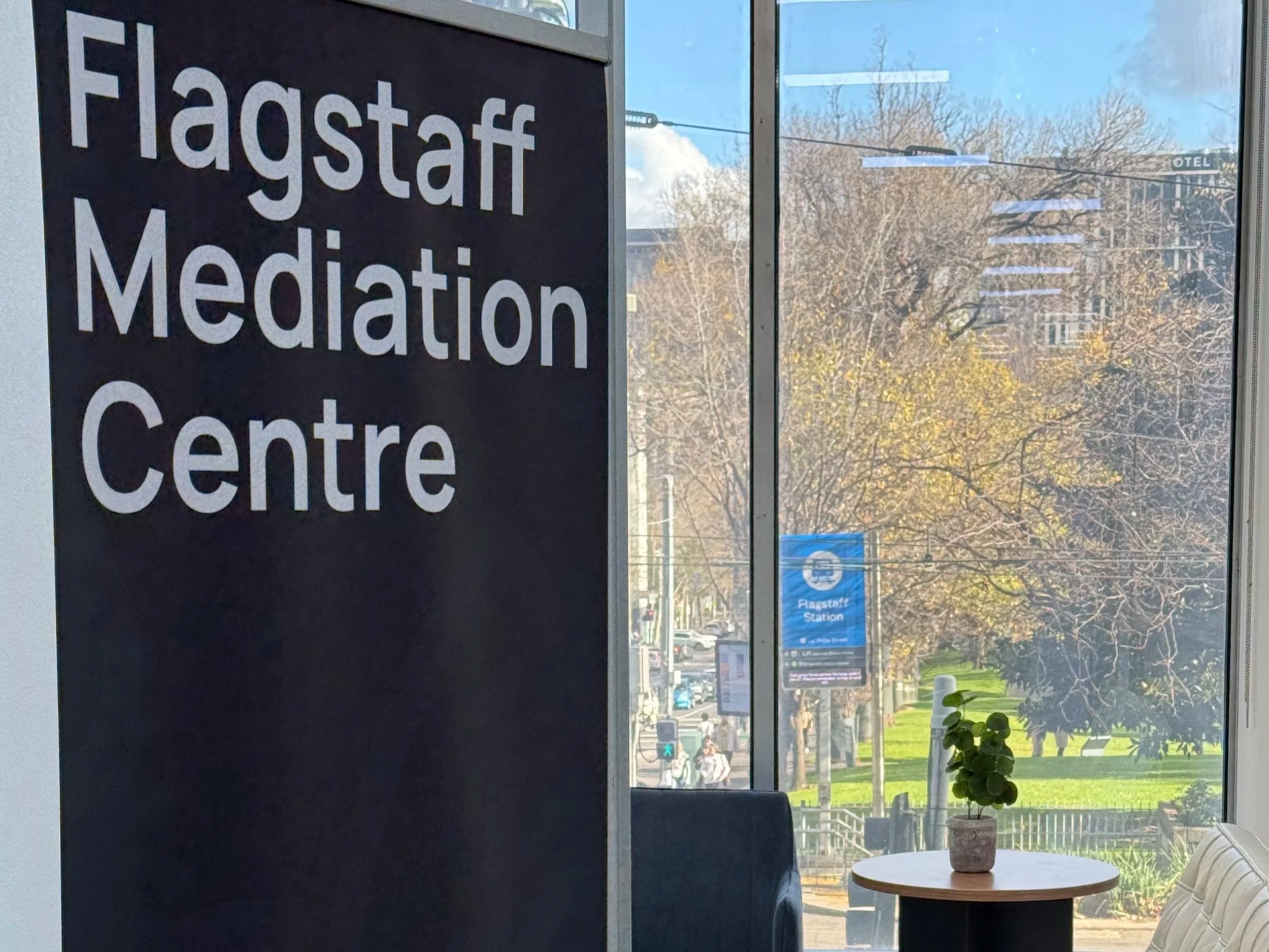 View from room inside Flagstaff mediation Centre, through a large window overlooking the Flagstaff Gardens trees and nearby buildings. Inside, a sign reads 'Flagstaff Meditation Centre' and a table with a potted plant is near the window.