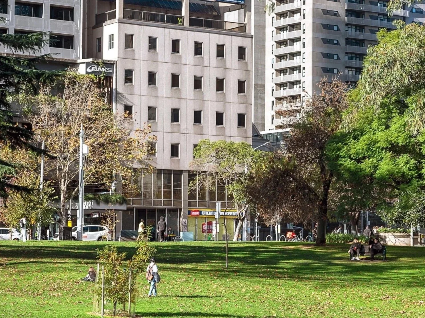 Flagstaff Mediation Centre Melbourne, view of large windows from Flagstaff Gardens, an urban park with trees, grass, and people sitting on benches or walking, surrounded by tall buildings and storefronts.