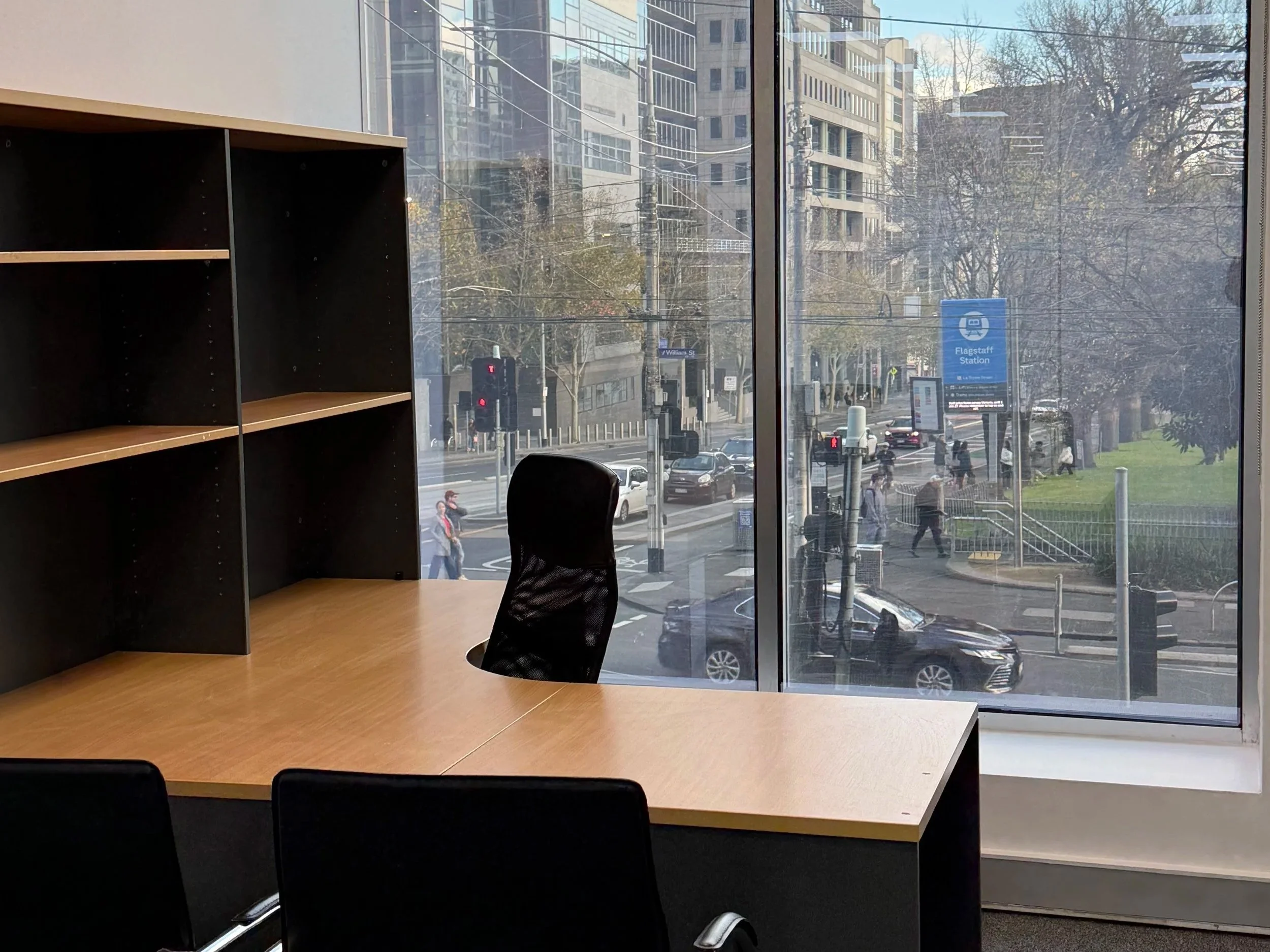 Flagstaff Mediation Centre Melbourne, view from inside a breakout room looking out large glass window showing city street with pedestrians, cars, and buildings, with a blue Flagstaff Station sign outside, and Flagstaff Gardens.