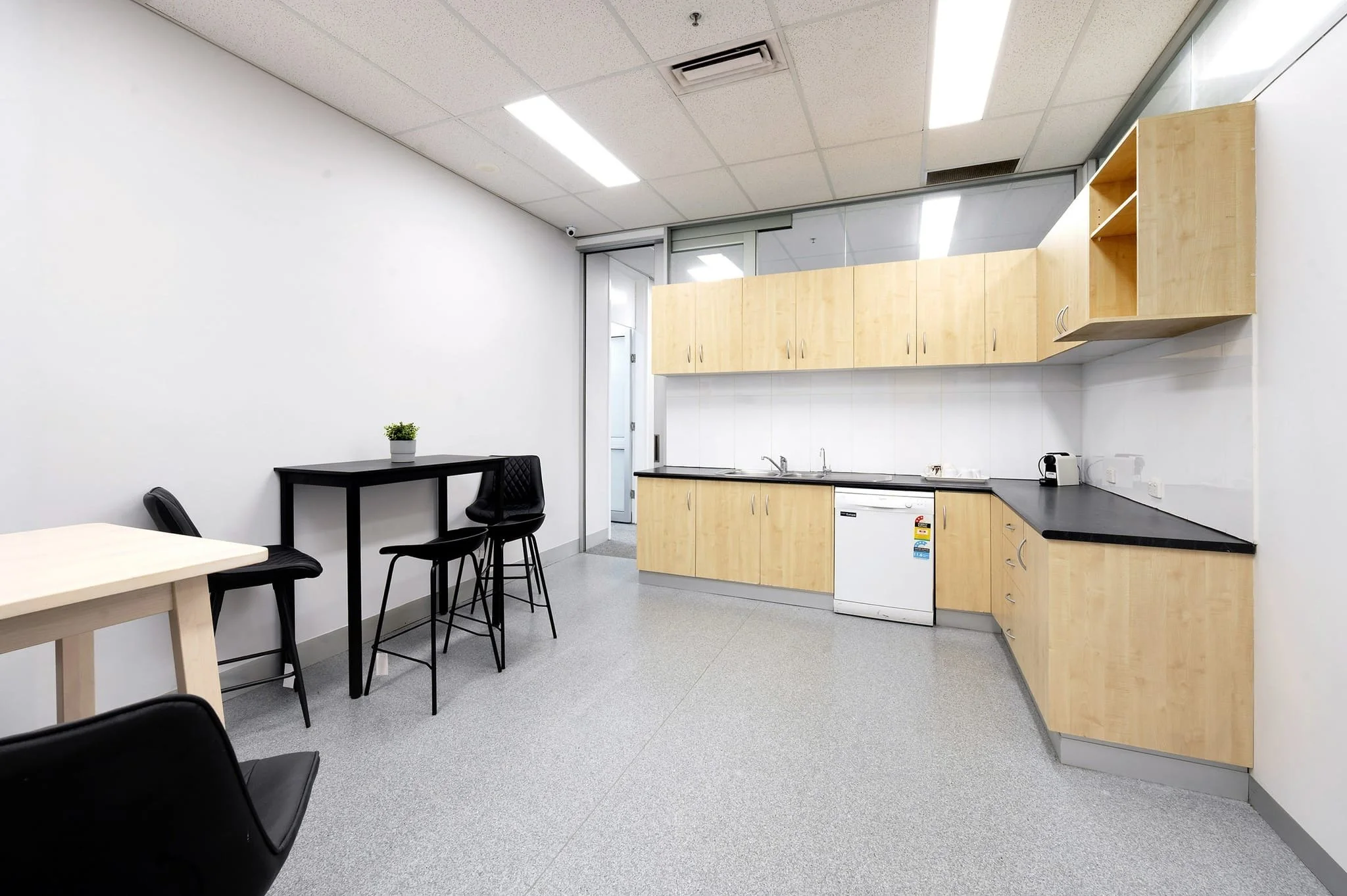 Flagstaff Mediation Centre kitchenette with wooden cabinets, a small refrigerator, a sink, a black countertop, a table with black chairs, and a small potted plant on the table. The room has white walls and a gray speckled floor with ceiling lights.