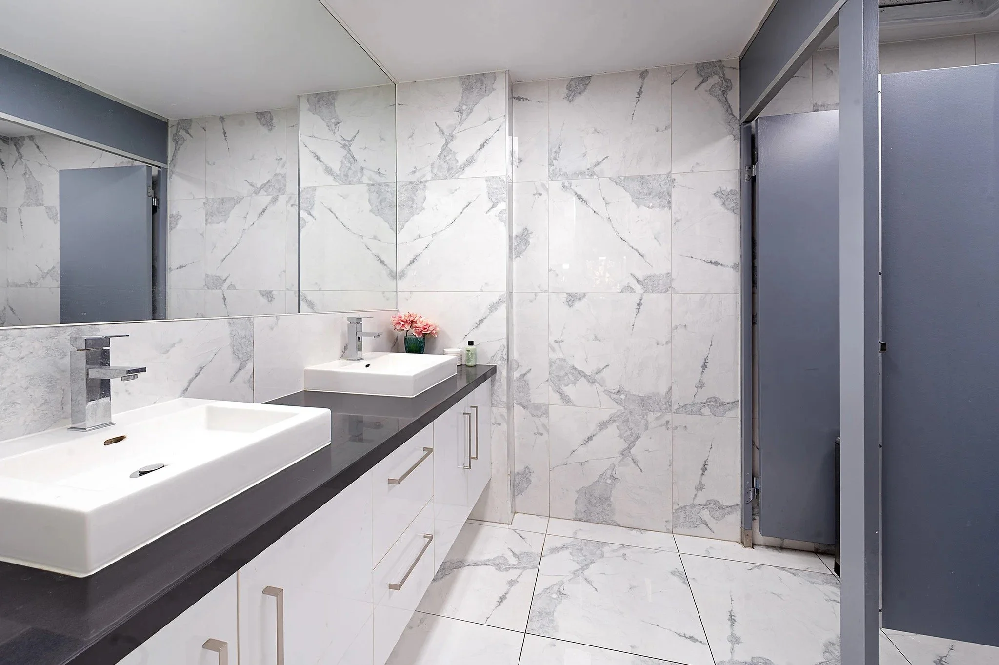 Flagstaff Mediation Centre Melbourne, bathroom with marble tiled walls, two rectangular vessel sinks on a gray countertop, a large mirror, flowers in a vase, and bathroom fixtures.