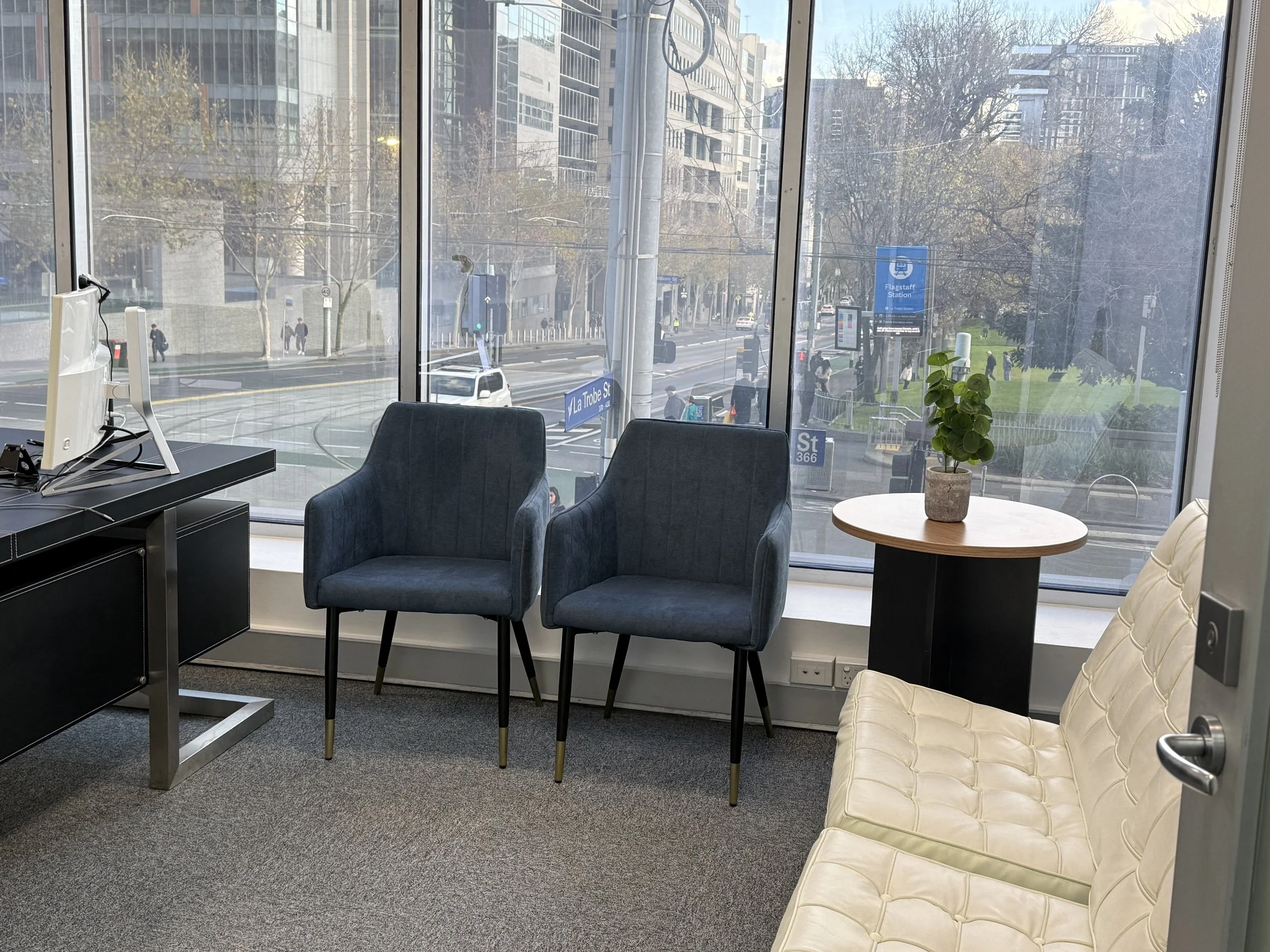 Flagstaff Mediation Centre Melbourne, view through large windows to Flagstaff Gardens trees, street signs, and buildings, taken from inside an office with chairs, a small table with a potted plant, and a computer on a desk.