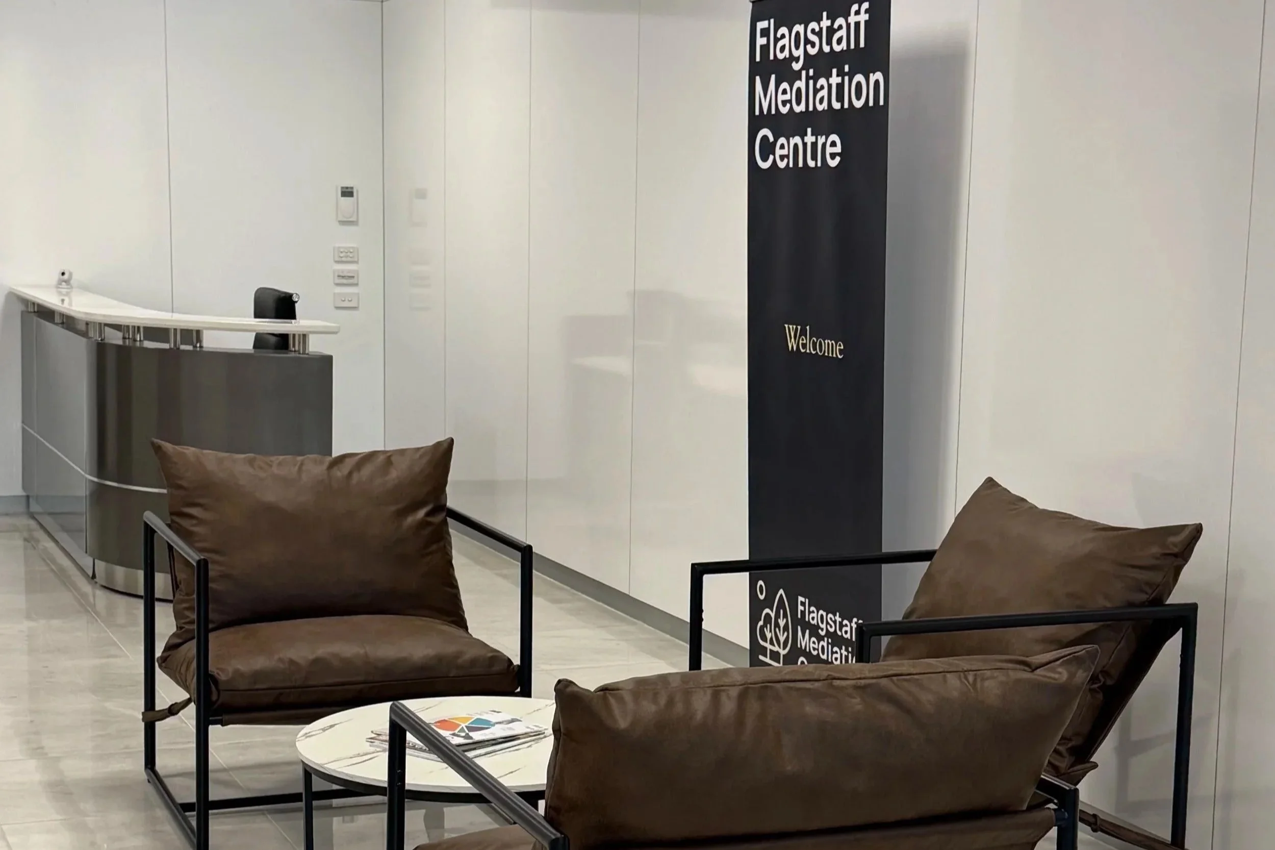 Interior of Flagstaff Mediation Centre Melbourne with main reception area, dark chairs with brown cushions, a white marble table, and a black sign with white text welcoming visitors.
