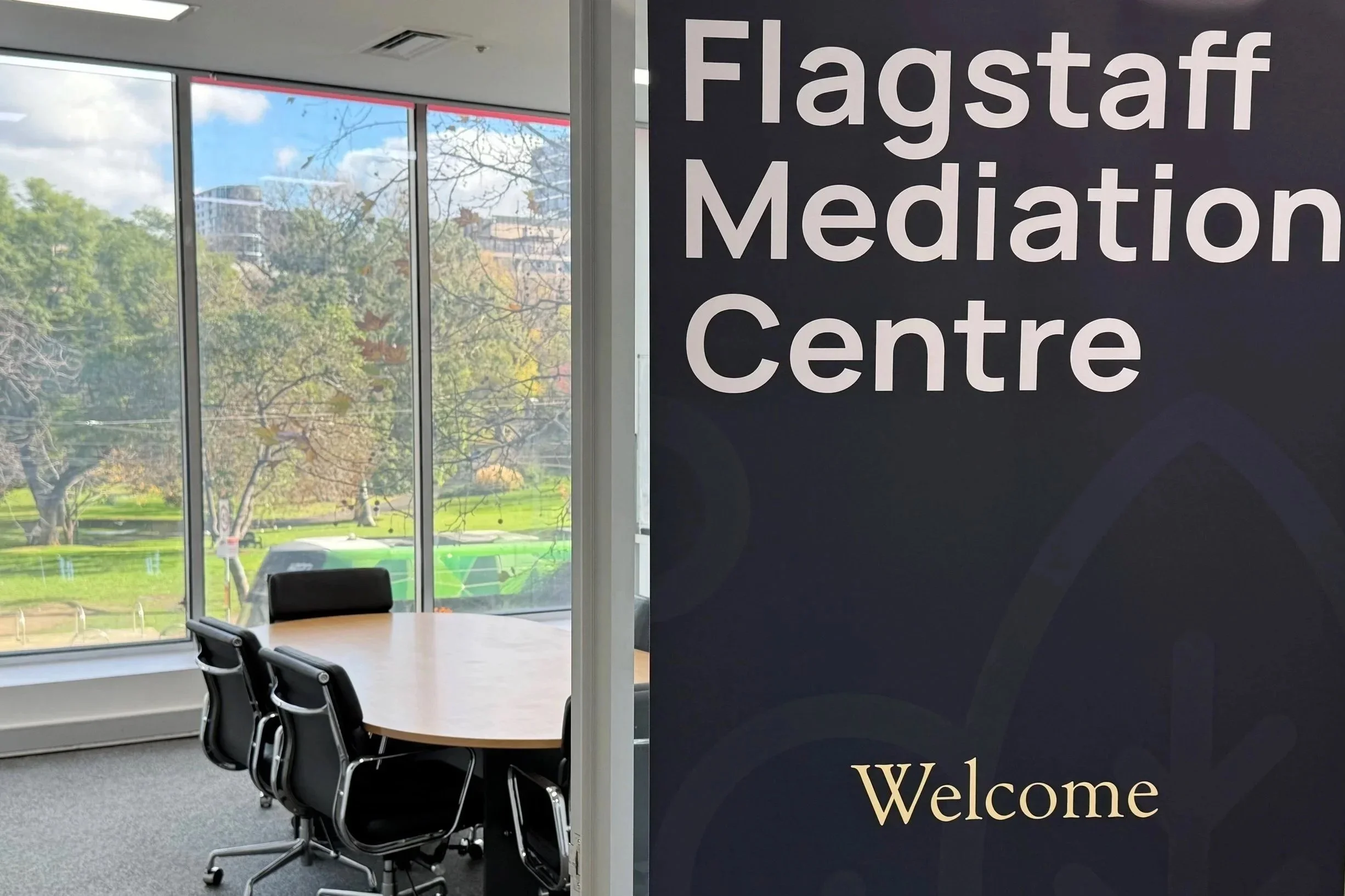 Flagstaff Mediation Centre Melbourne, view through large windows to Flagstaff Gardens trees and buildings outside. A dark sign with the words "Flagstaff Meditation Centre" and "Welcome" partially covers the room.
