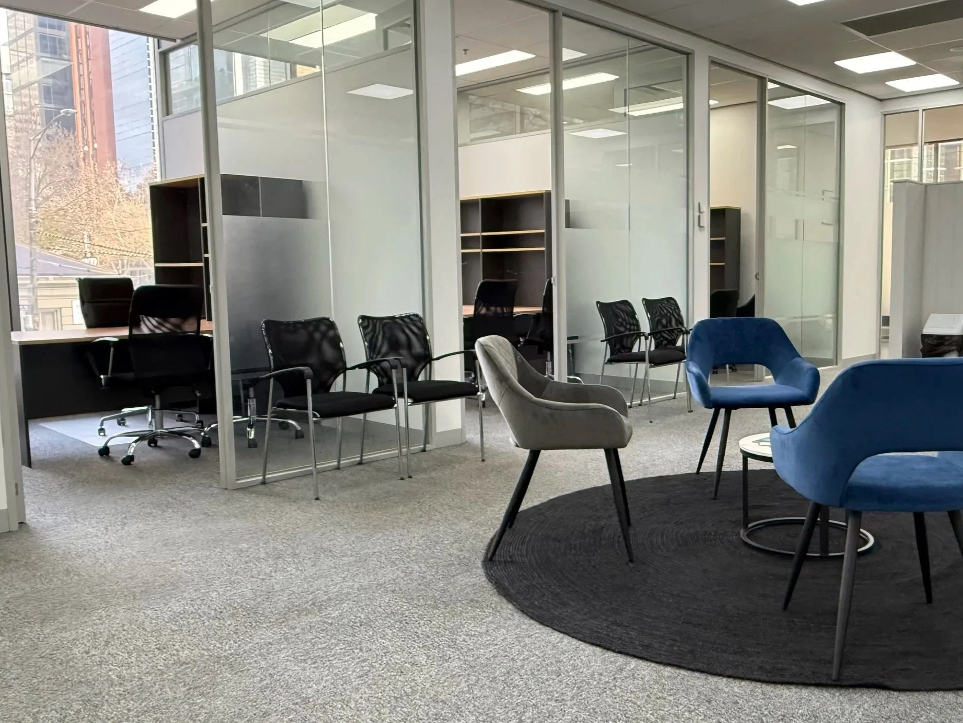 Flagstaff Mediation Centre second reception area, a modern office space with glass-walled breakout meeting rooms, black and grey lounge chairs, chairs along the walls, and a building with trees outside visible through large windows.