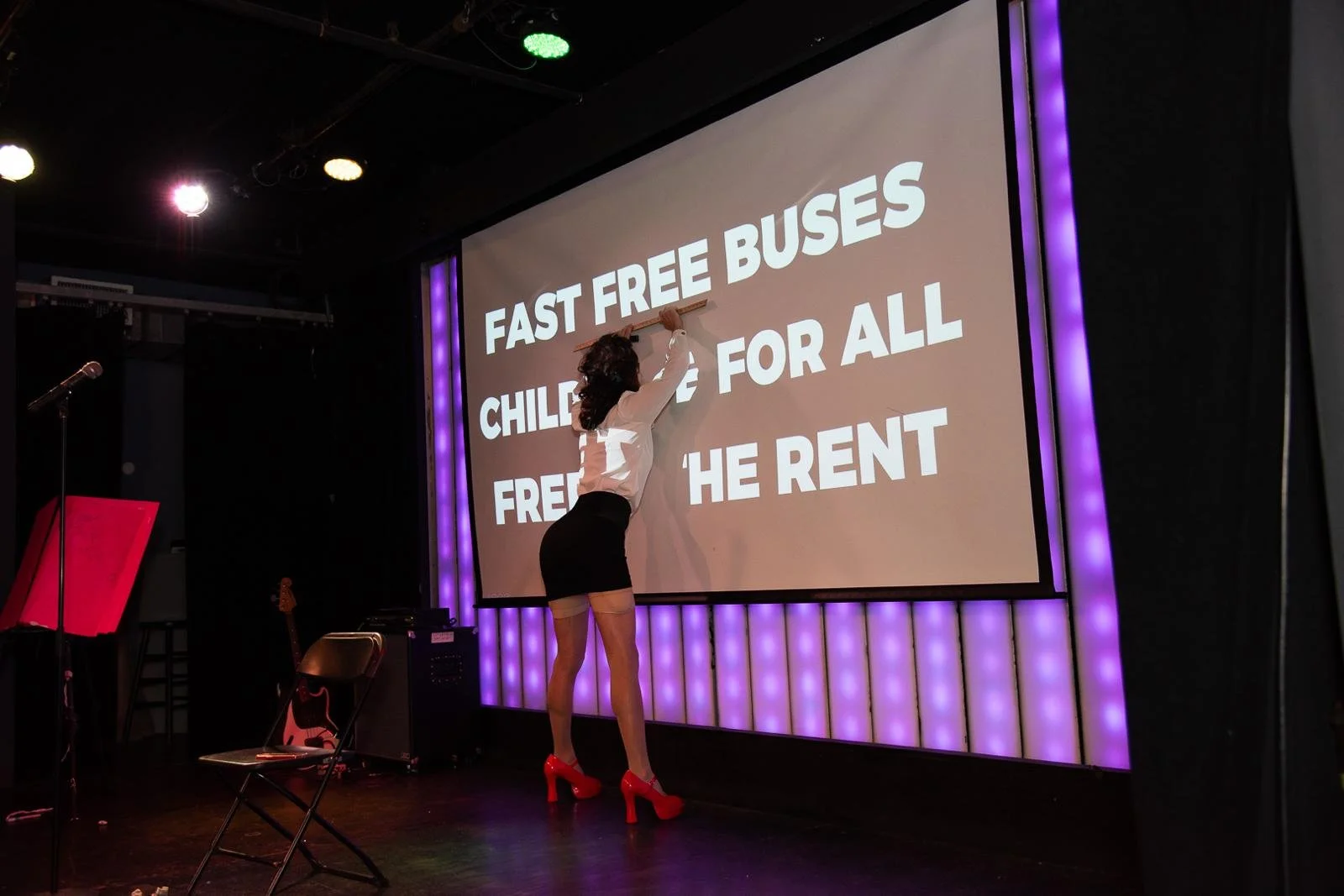 A woman writing on a presentation screen that reads 'Fast free buses children for all free the rent' on a stage with purple and green lighting.