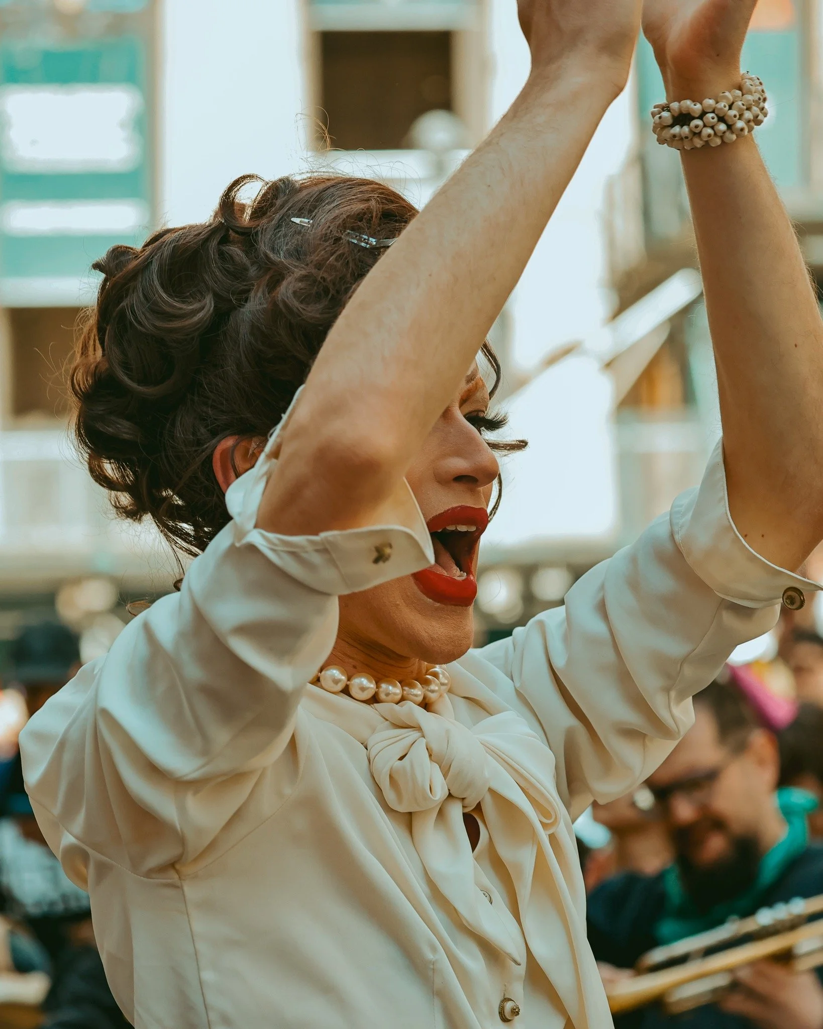 The drag queen Monaschewitz is at a May Day protest. Her arms are raised, blocking her face. She wears pearls and red lipstick. Photo taken by Framed Unrest.