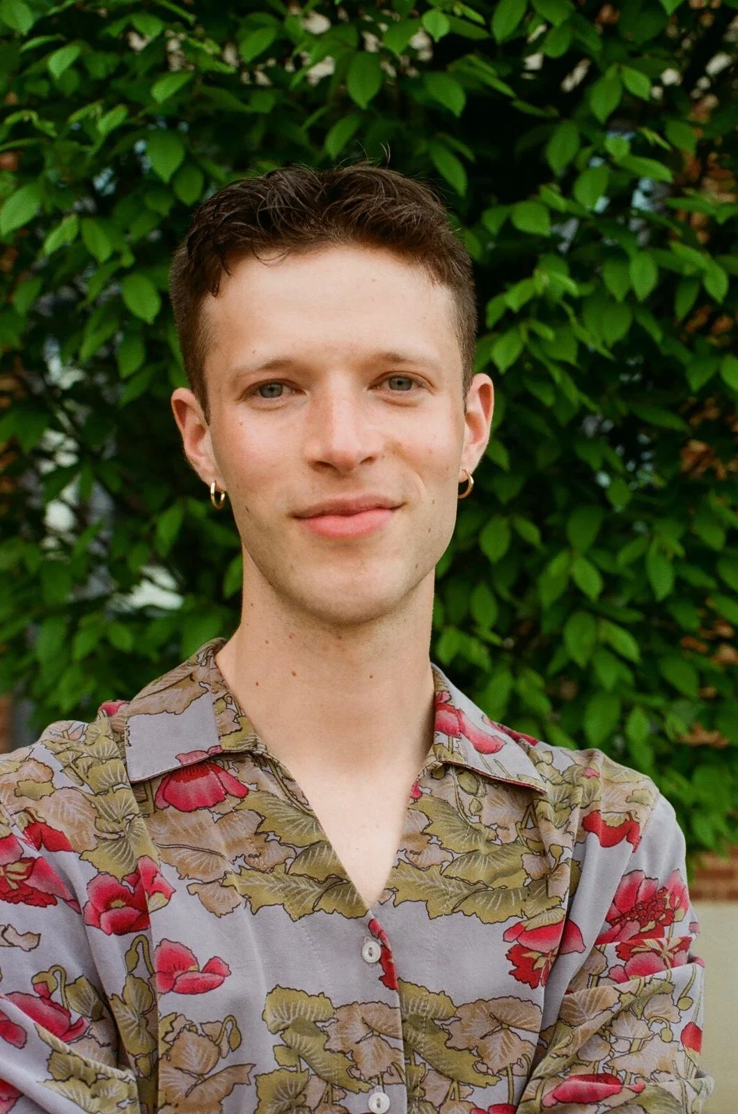 Isaiah stands in front of a leafy bush, wearing a lavender shirt with red poppies on it. Isaiah has small gold hoop earrings and smiles slightly.