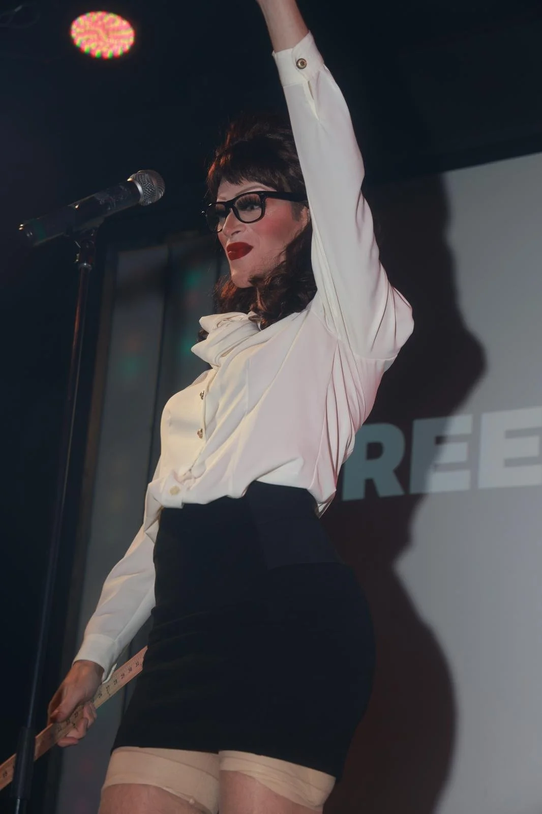 The drag queen Mona Schewitz stands on a stage in a black skirt, a white blouse, and a wig with bang. She holds a yard stick.