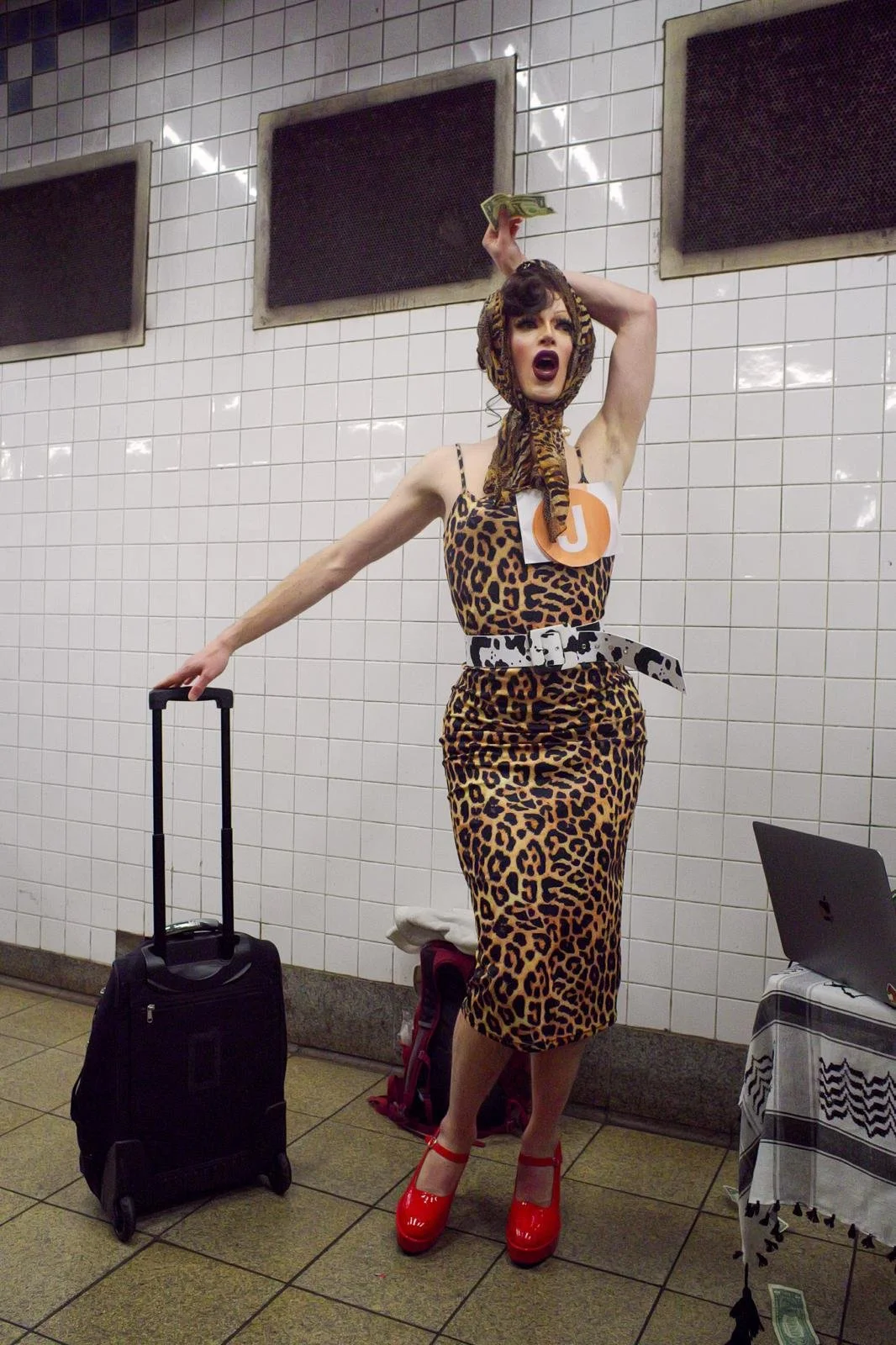 The drag queen Mona Schewitz stands in a subway station wearing a leopard print dress and red heels. She holds a suitcase with one  hand, the other hand is outstretched above her head holding a dollar bill. Photo taken by Dill Dough King